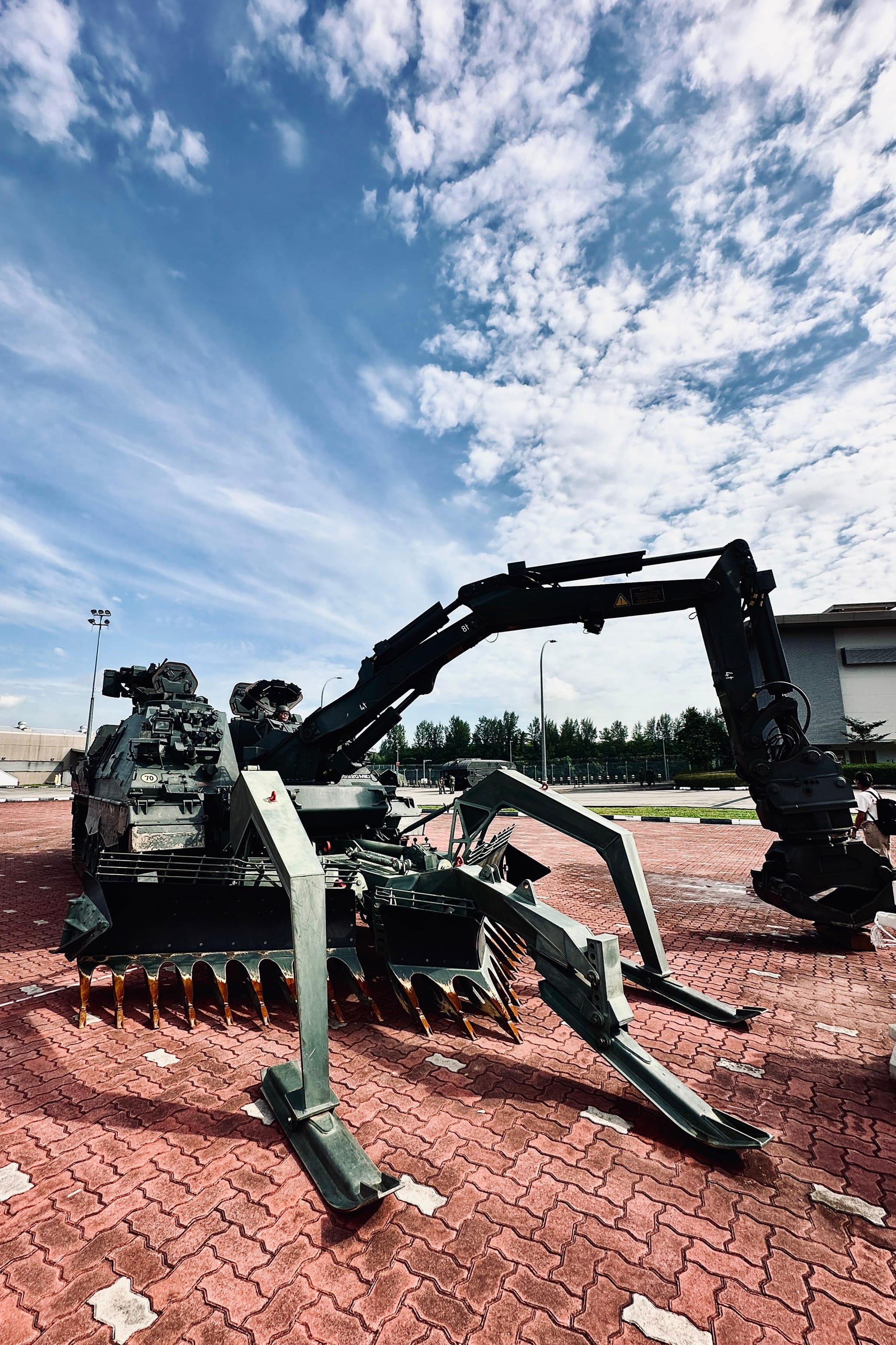 A large black military vehicle with a mine flail and mechanical arm sits on a red brick surface under a blue sky.