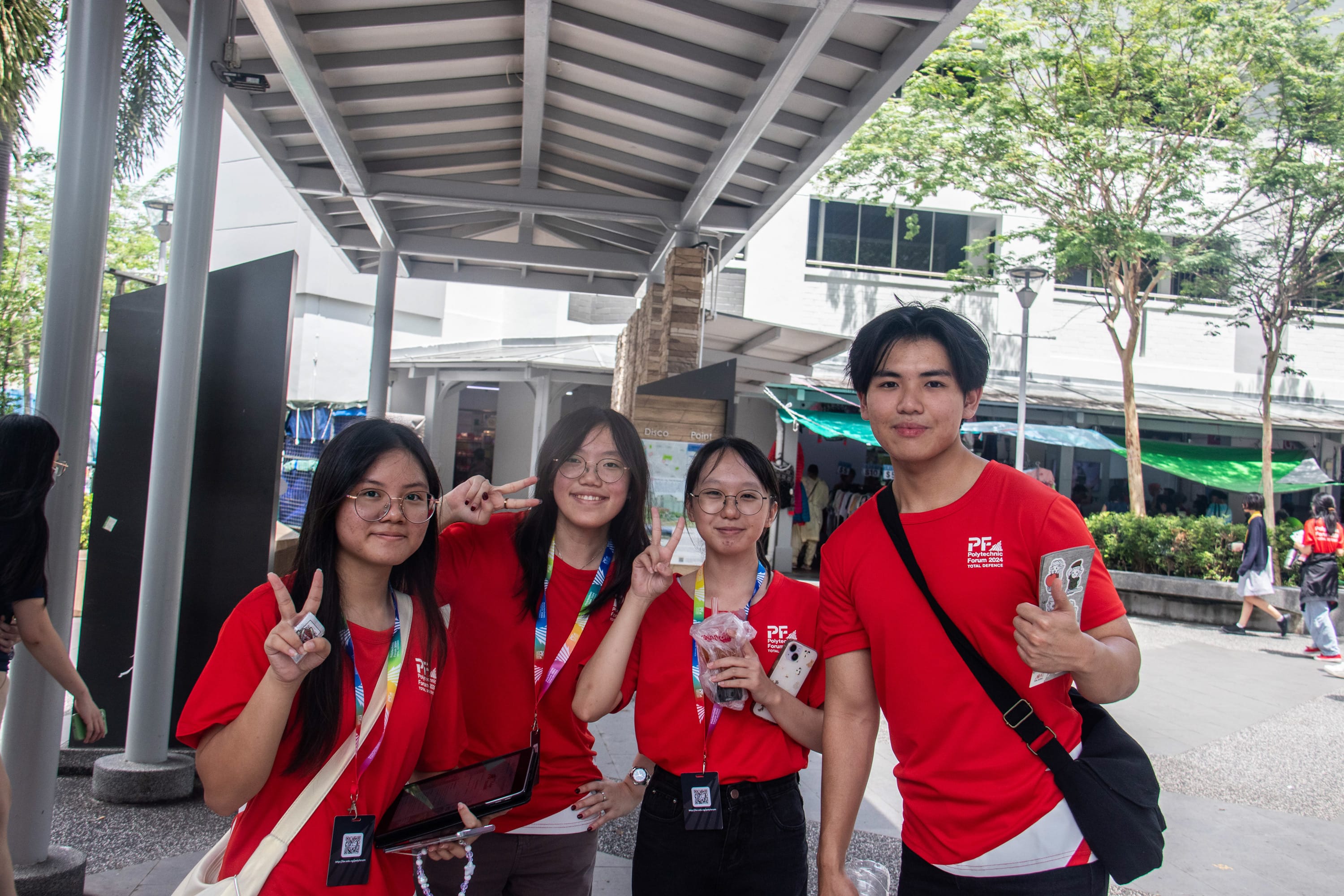Four young people in matching red Polytechnic Forum shirts and lanyards smile and pose outdoors under a covered walkway.