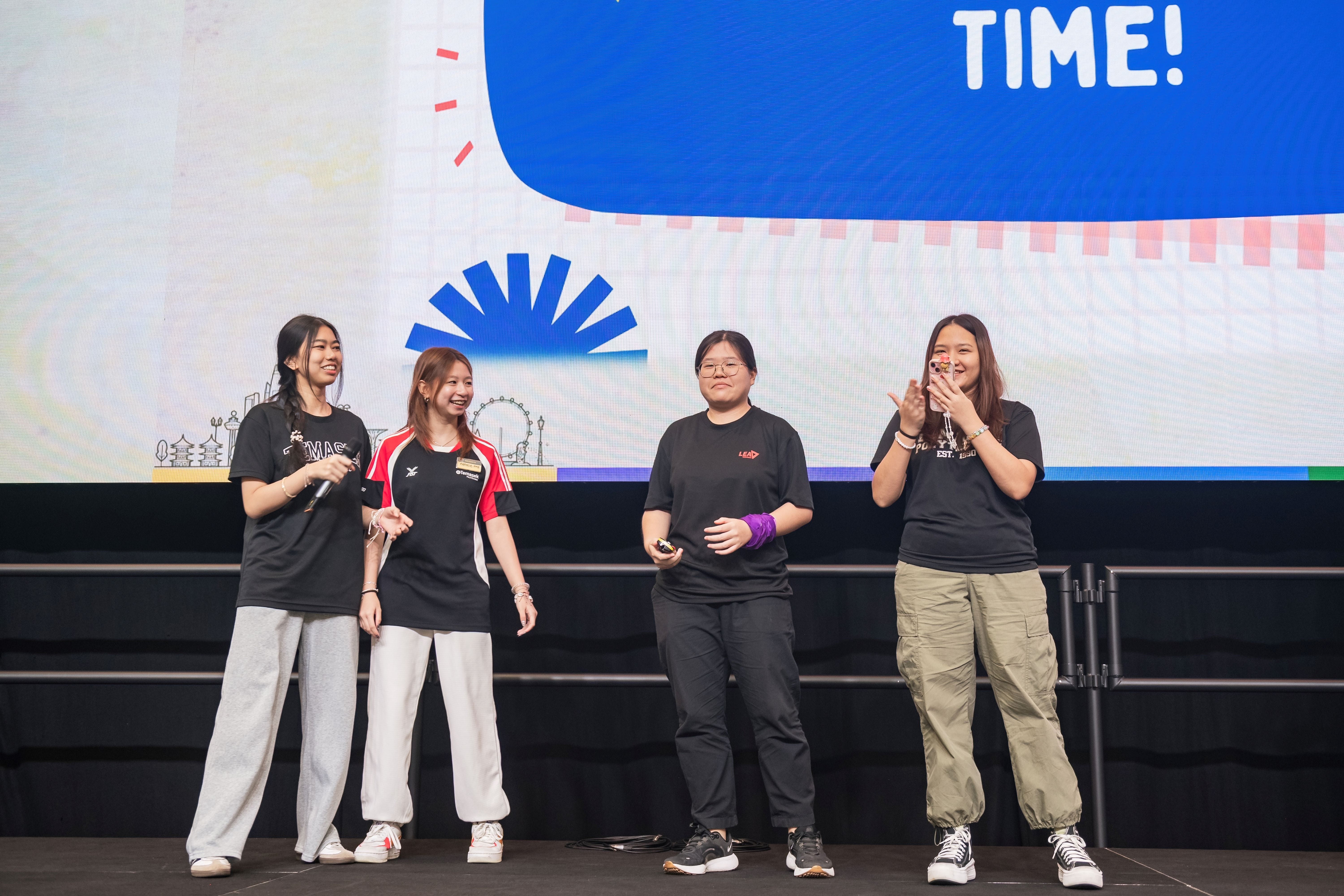 Four young women stand on a stage smiling, with a blue screen displaying the word TIME behind them.