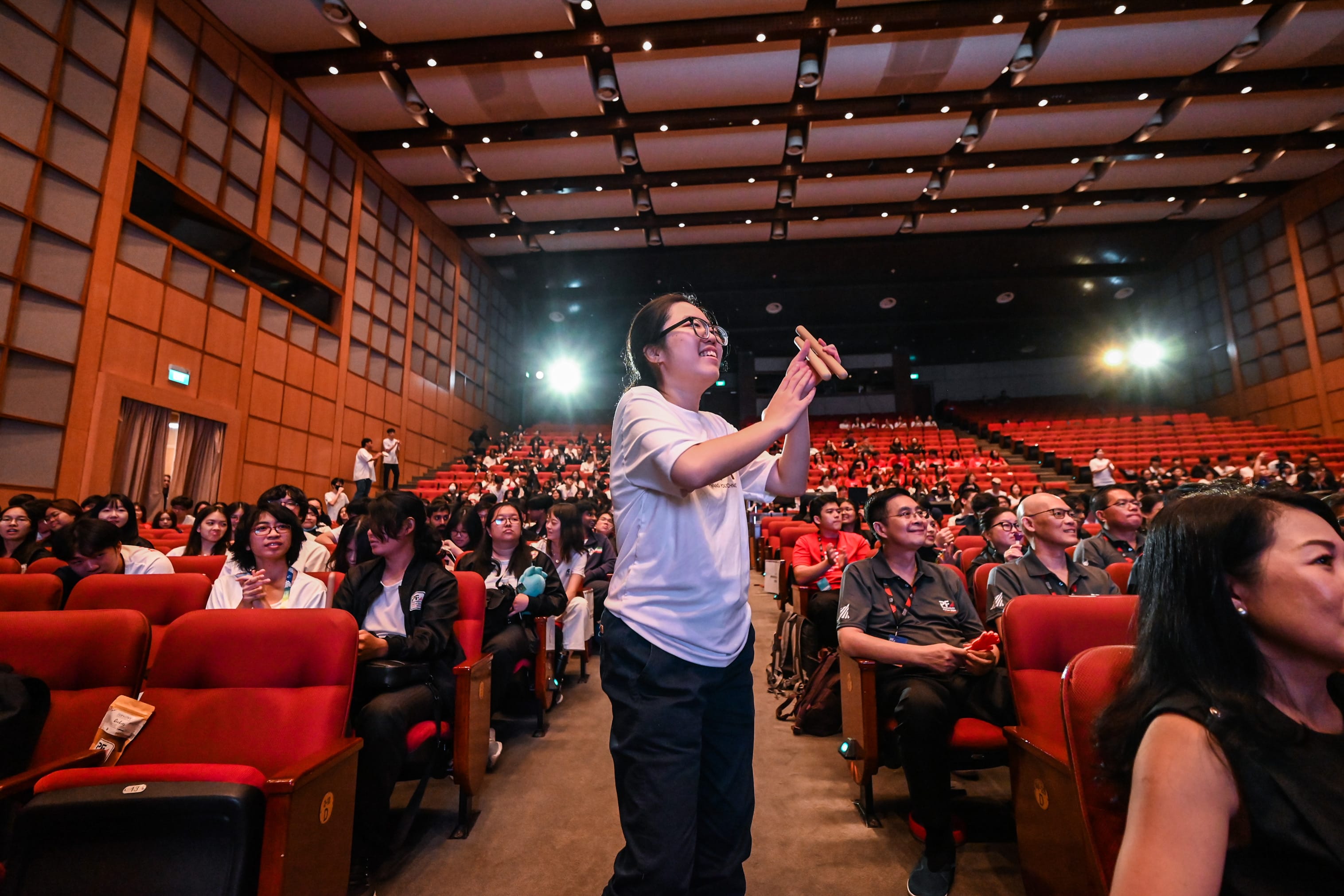 A smiling young woman in a white shirt stands in a packed auditorium with red seats, holding an object up.