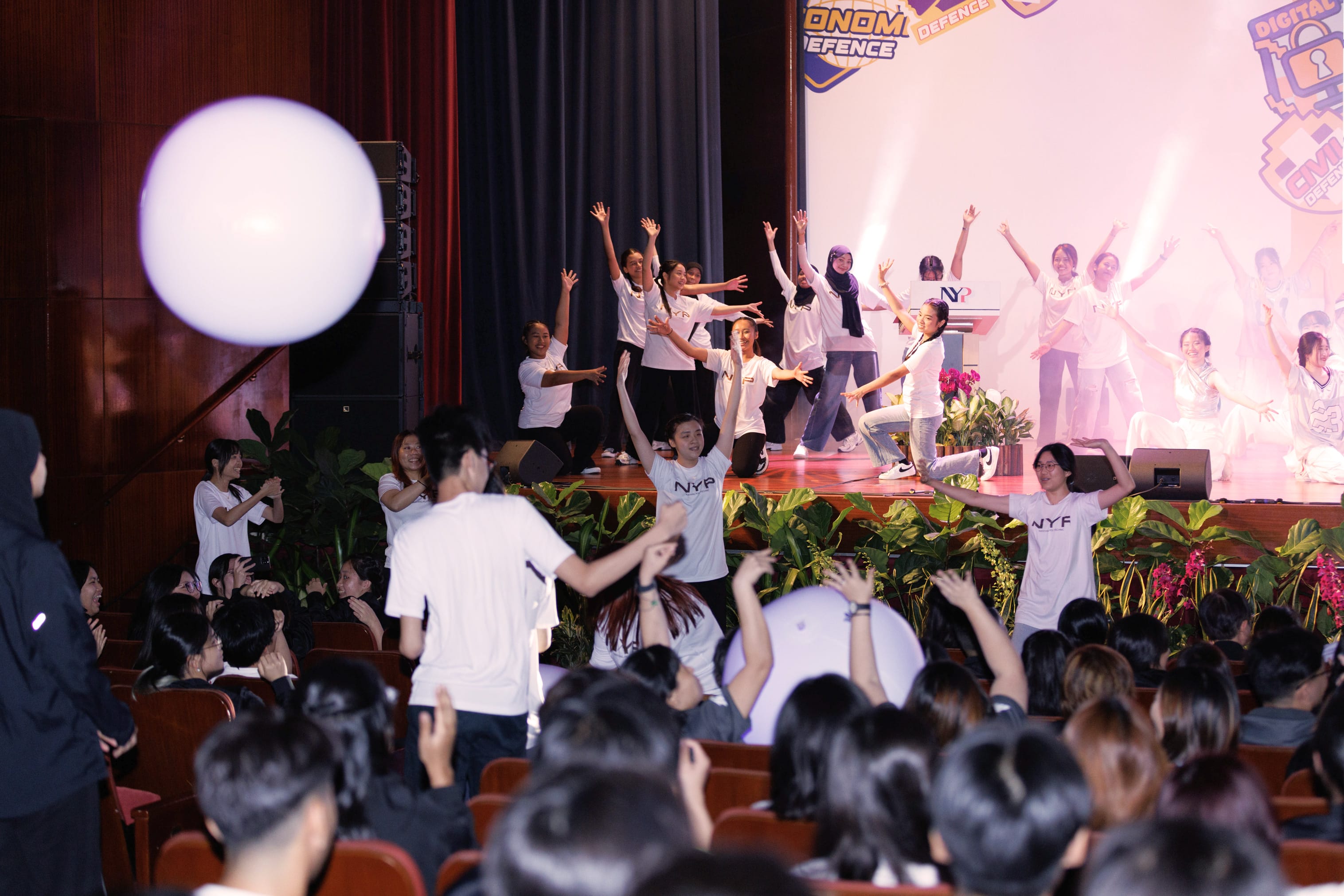 Students in white NYP shirts dance on and below a stage before a seated audience, with a white balloon in the foreground.