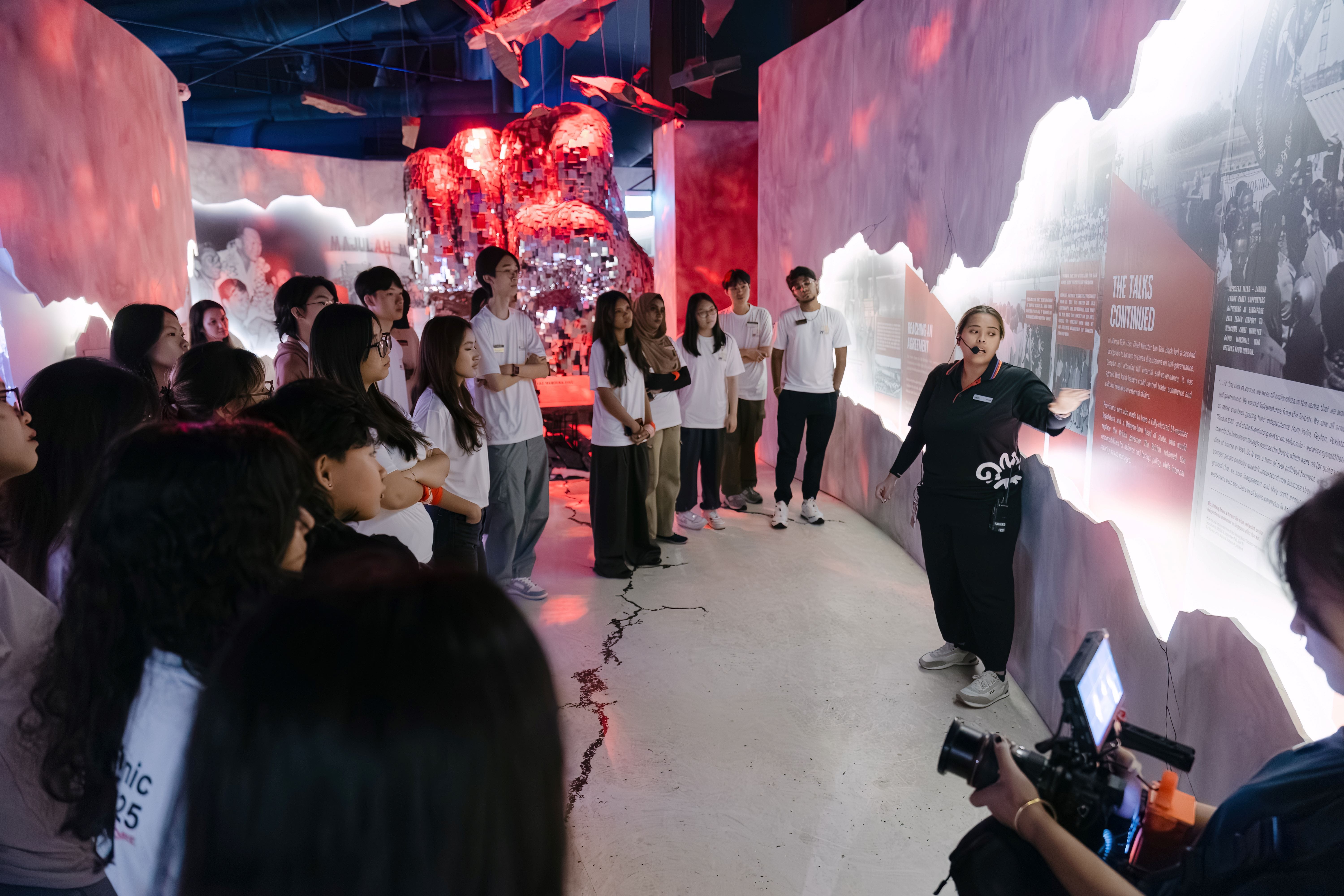 A guide in black speaks to a group of young visitors in a dimly lit museum exhibit with red sculptural installations.