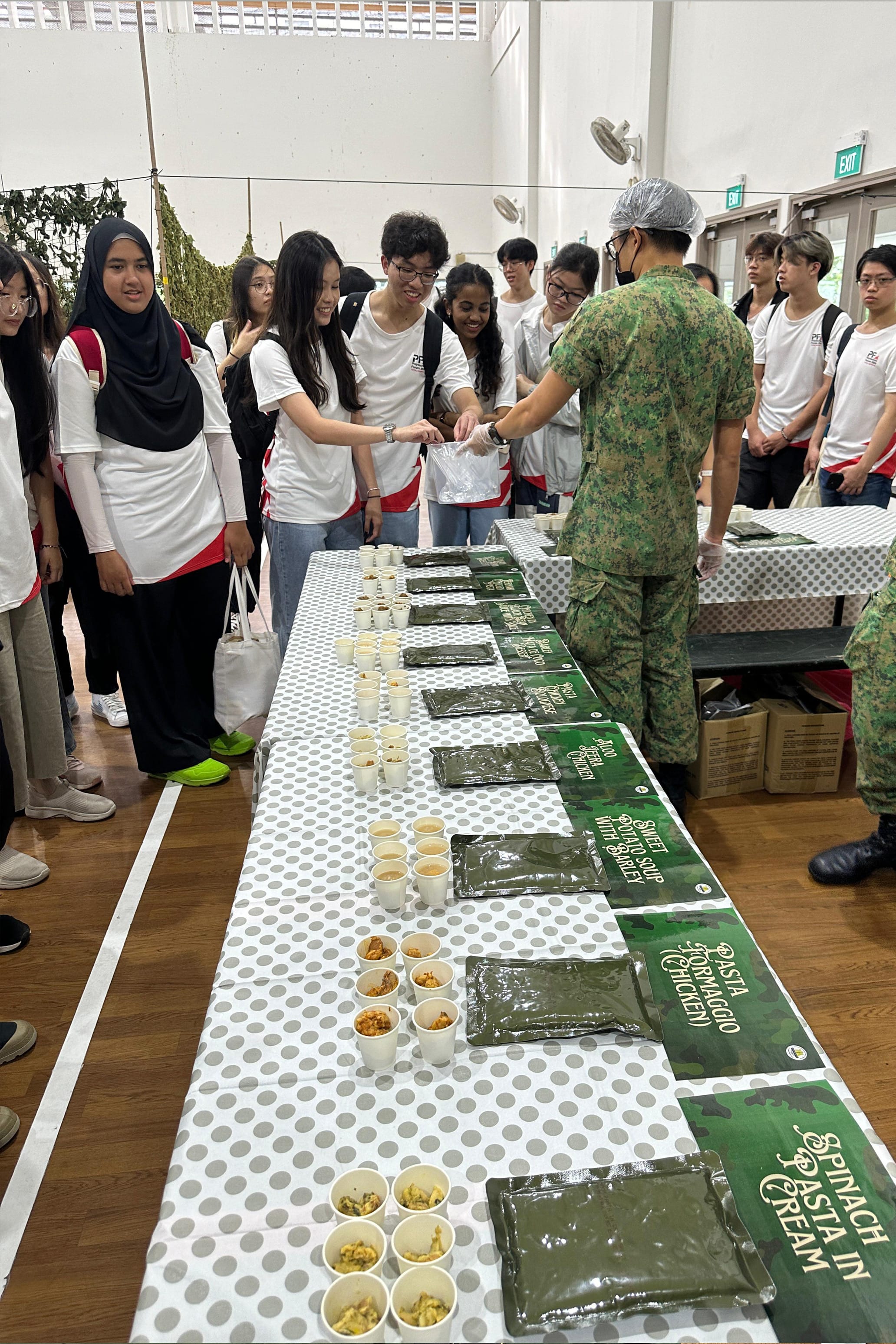 Students in white shirts gather around a long table with military ration packs and small cups of food samples.