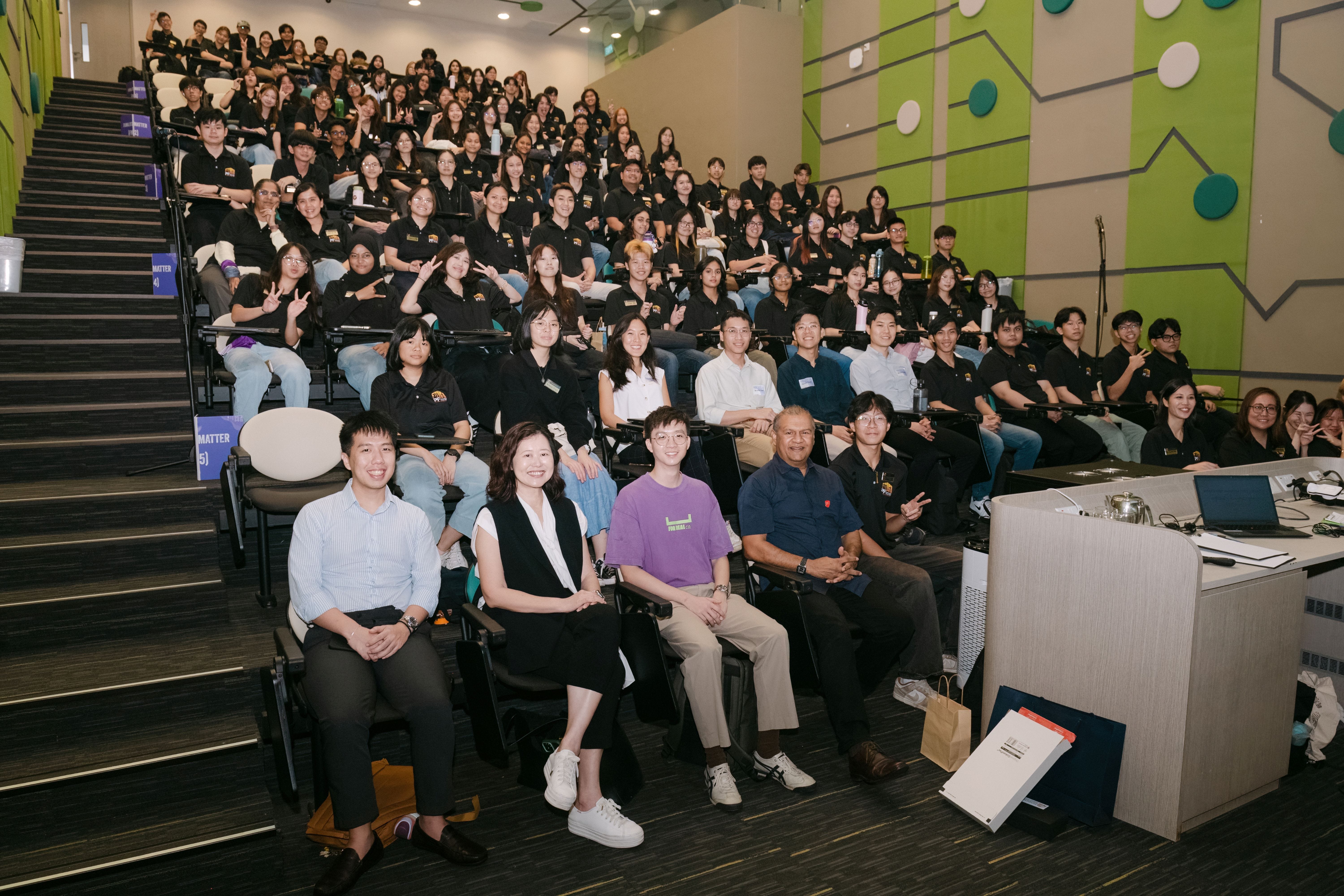 Large group of students in matching black shirts filling a tiered lecture hall, posing for a photo with staff in front.