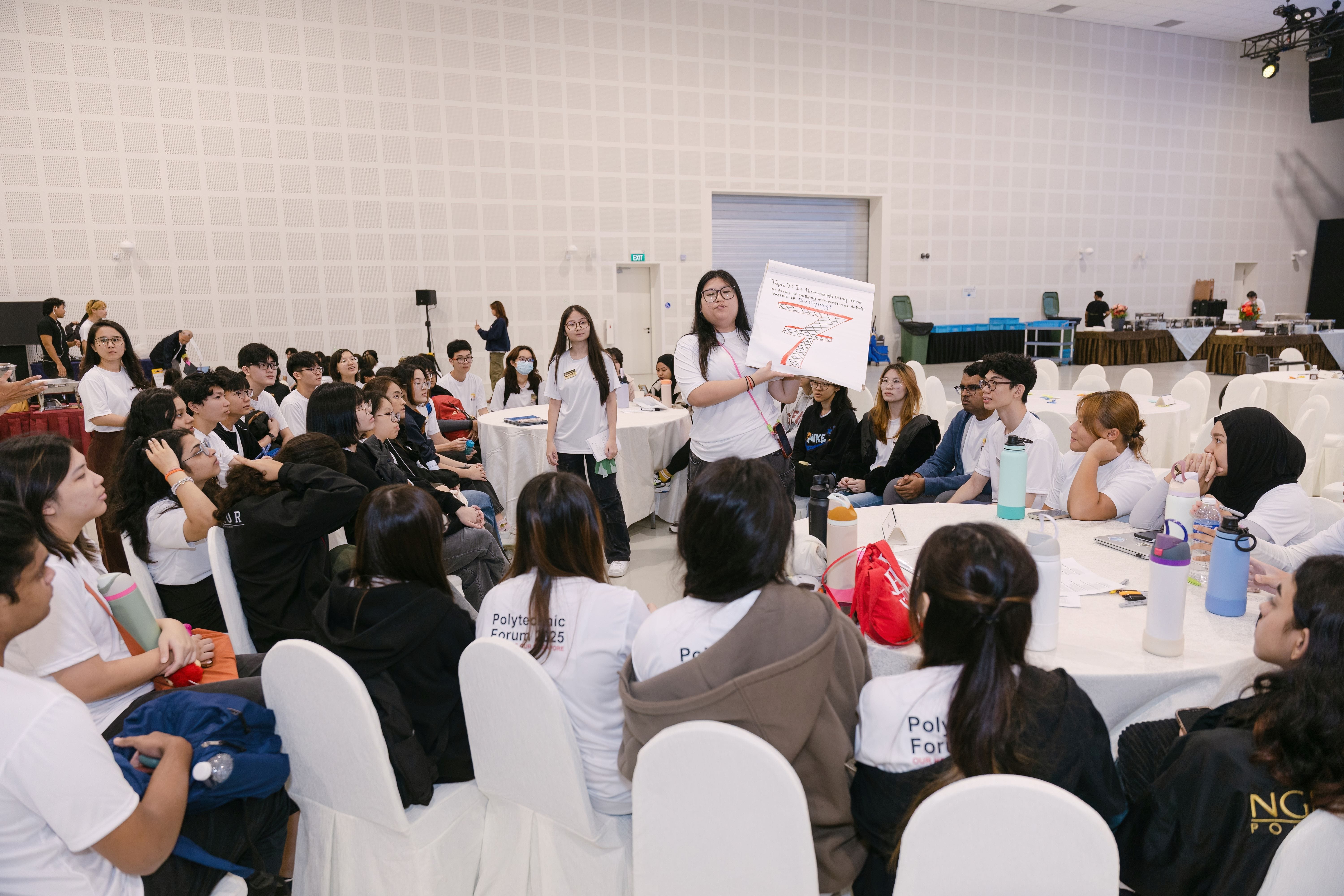 A young woman holds up a poster while presenting to a large group of students seated at round tables in a hall.