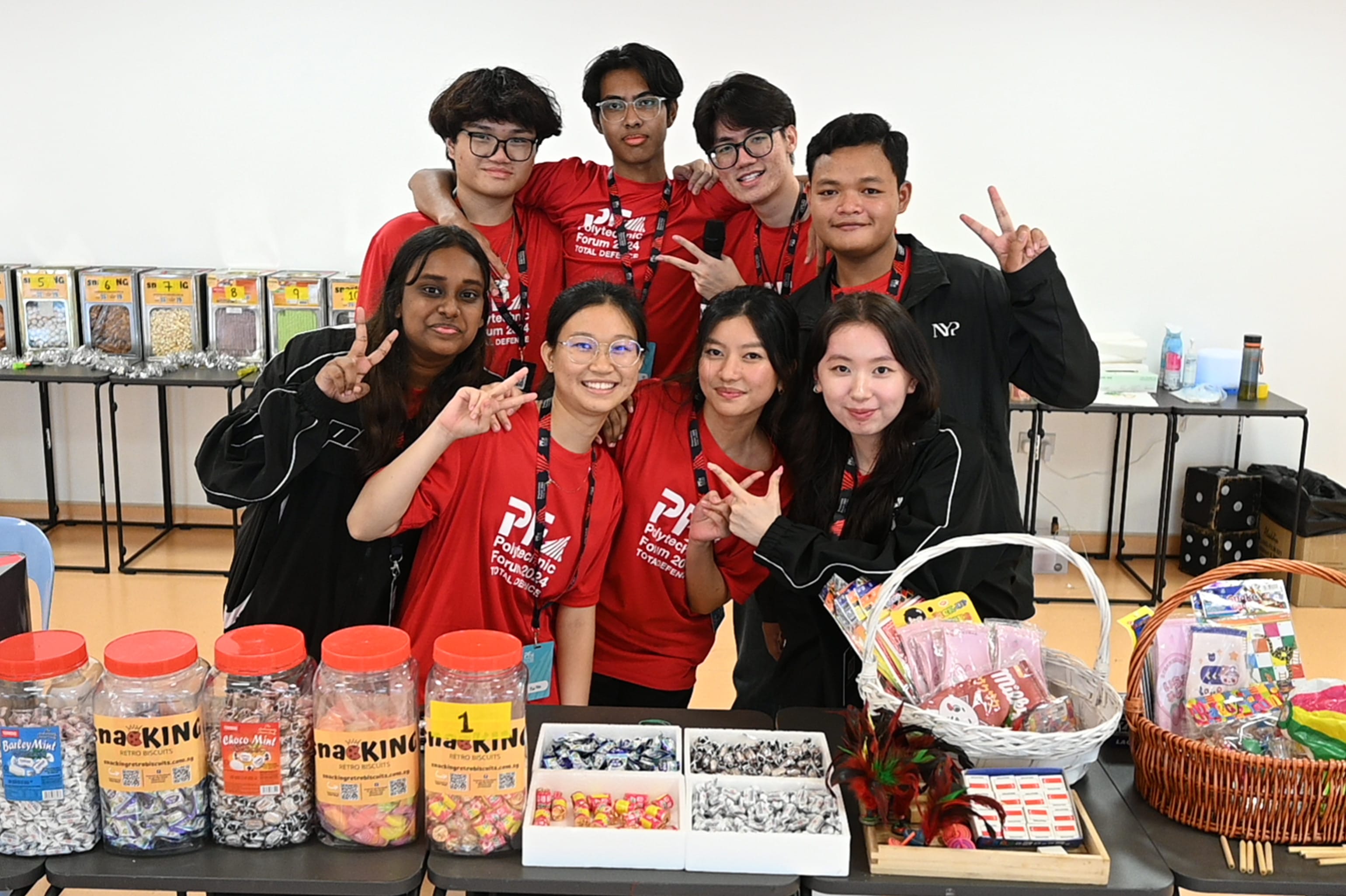 Eight smiling students in red Polytechnic Forum shirts pose behind a table of candy jars and snack baskets.