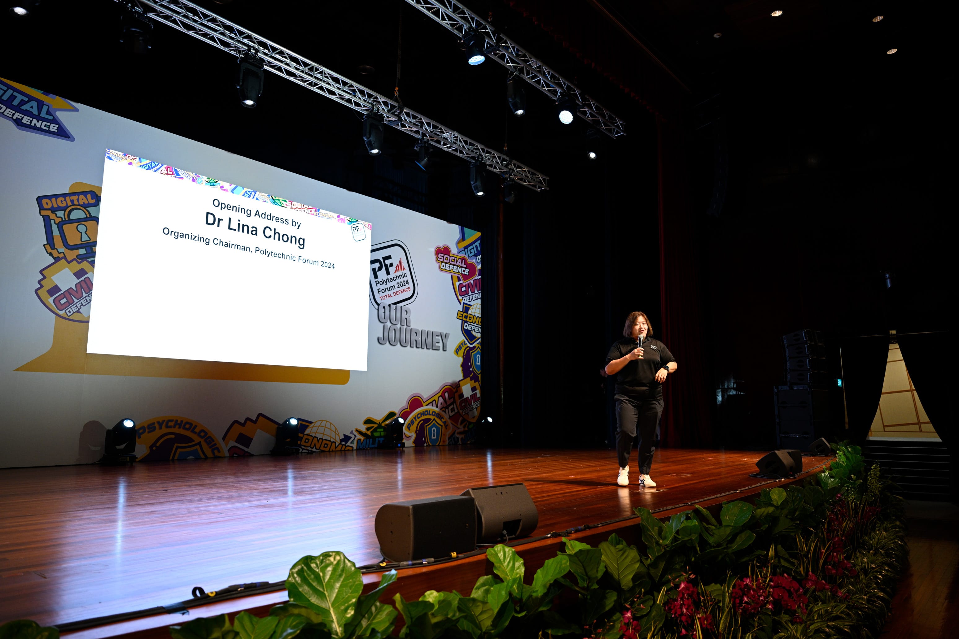 Dr Lina Chong holds a microphone on stage at Polytechnic Forum 2024, with a projected opening address slide behind her.