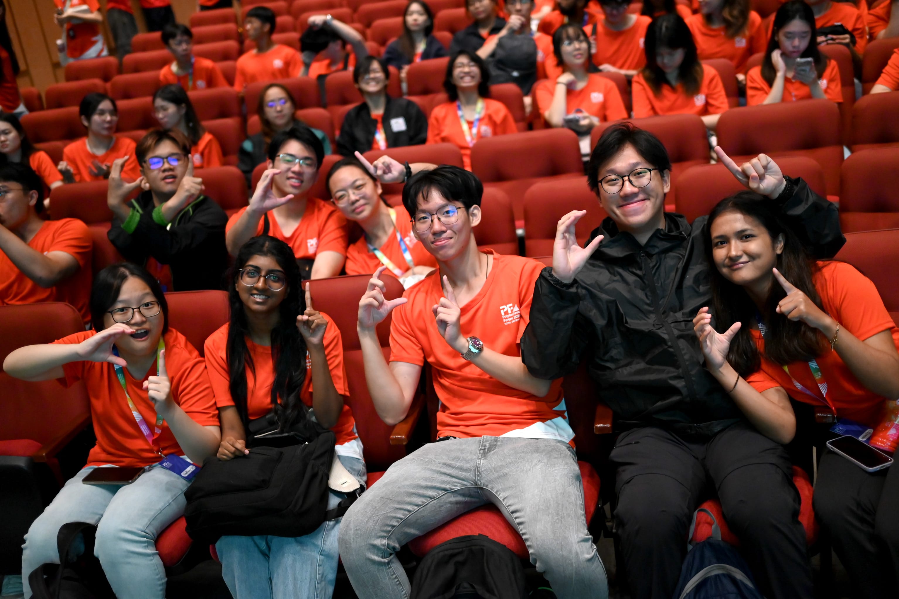 A large group of young people wearing orange shirts sit in a red auditorium, smiling and posing with hand gestures.