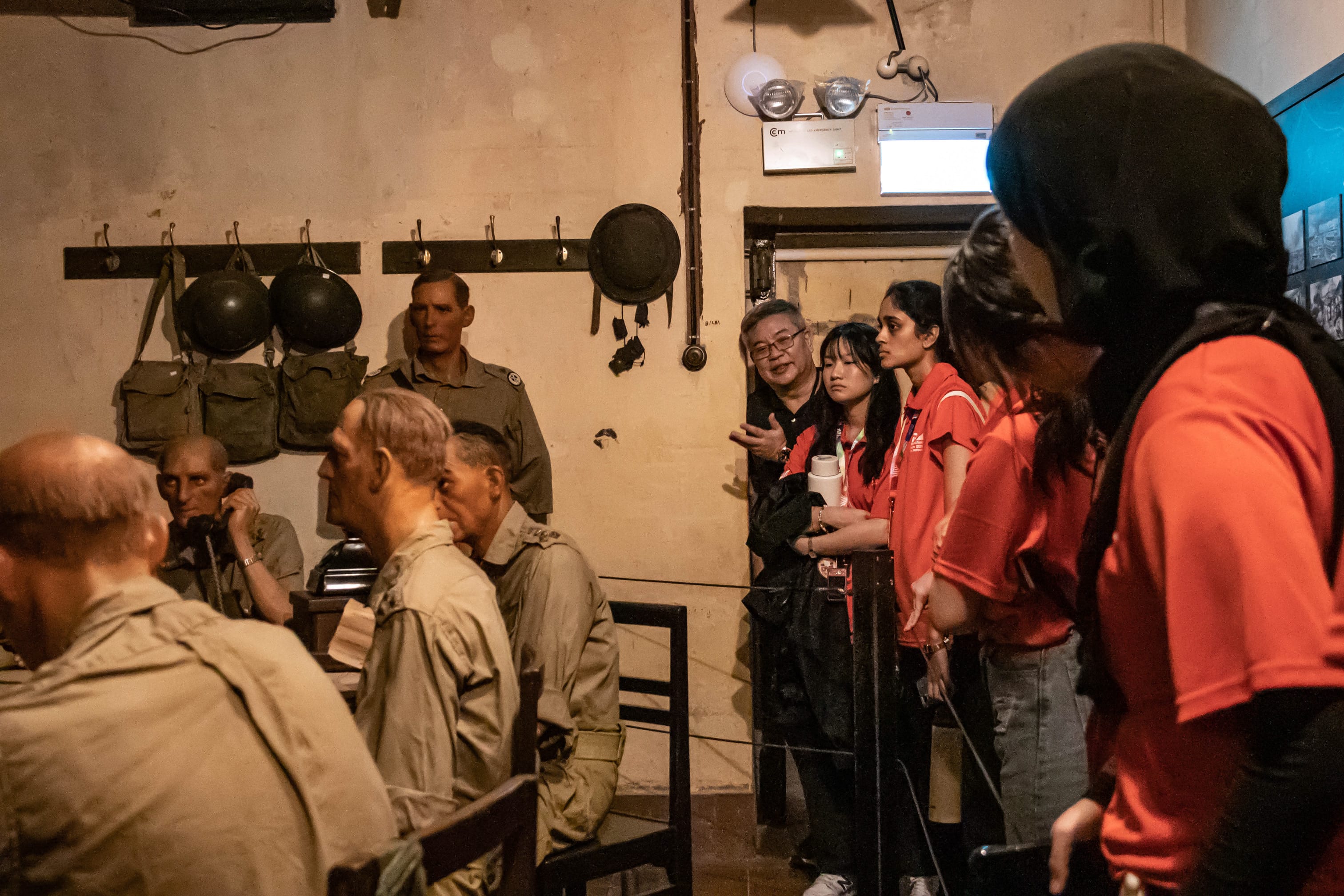 Visitors in red shirts observe wax figures of soldiers in military uniforms inside a dimly lit museum room.