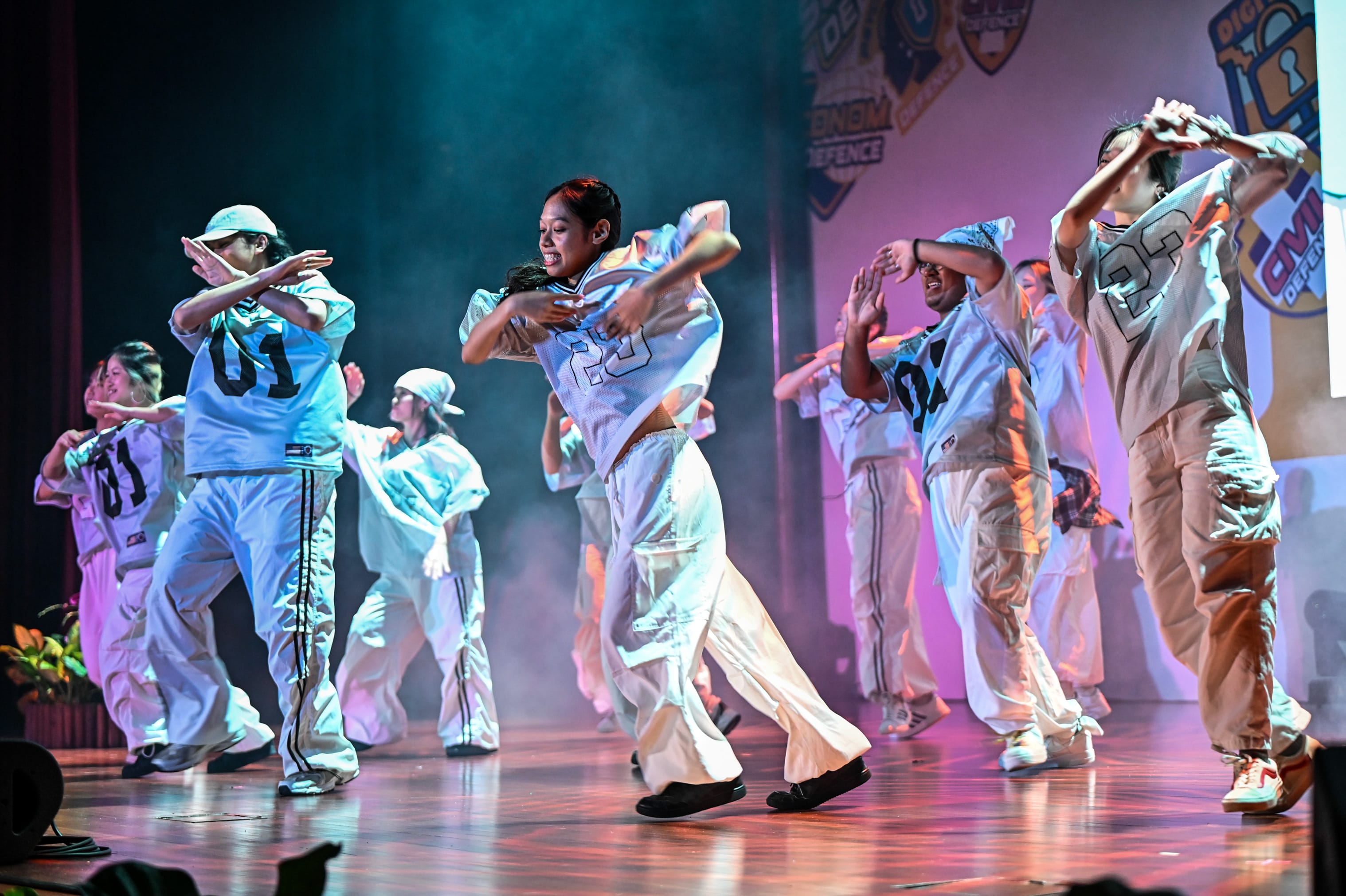 A group of dancers in numbered white jerseys and baggy pants perform hip-hop on a lit stage.