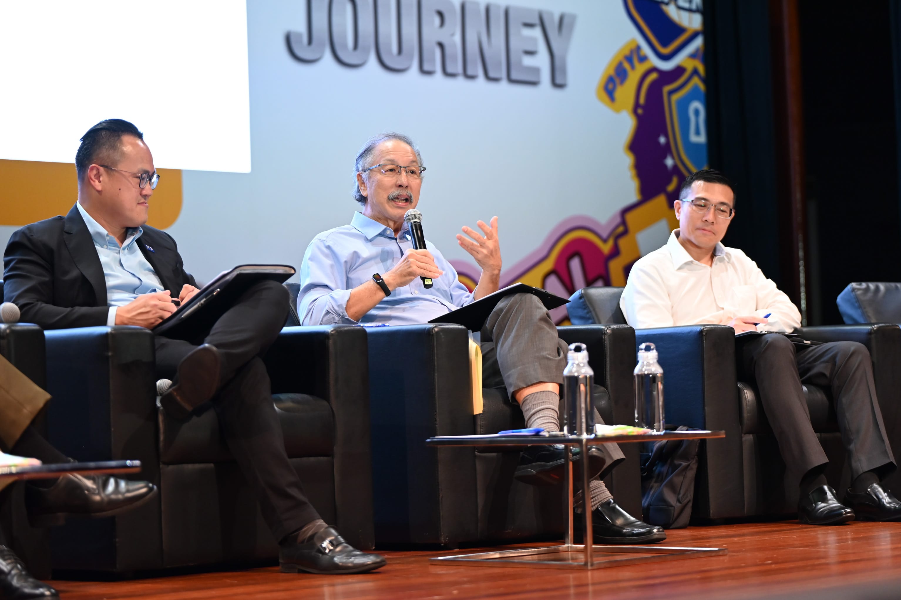 Three men sit on a panel stage, with the center man speaking into a microphone under a banner reading Journey.