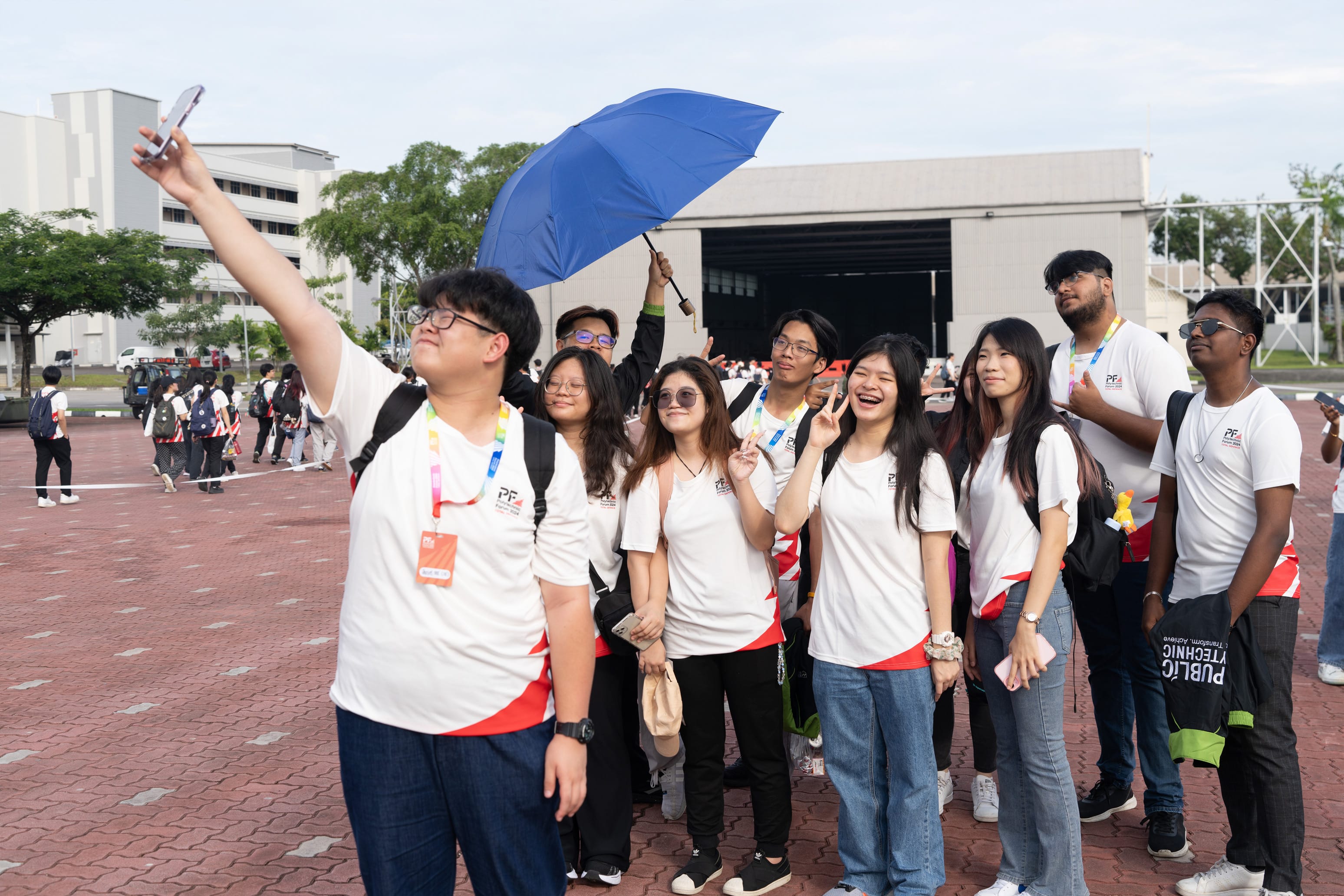 A group of young people in white shirts pose for a selfie outdoors, one holding a blue umbrella, smiling and making peace signs.