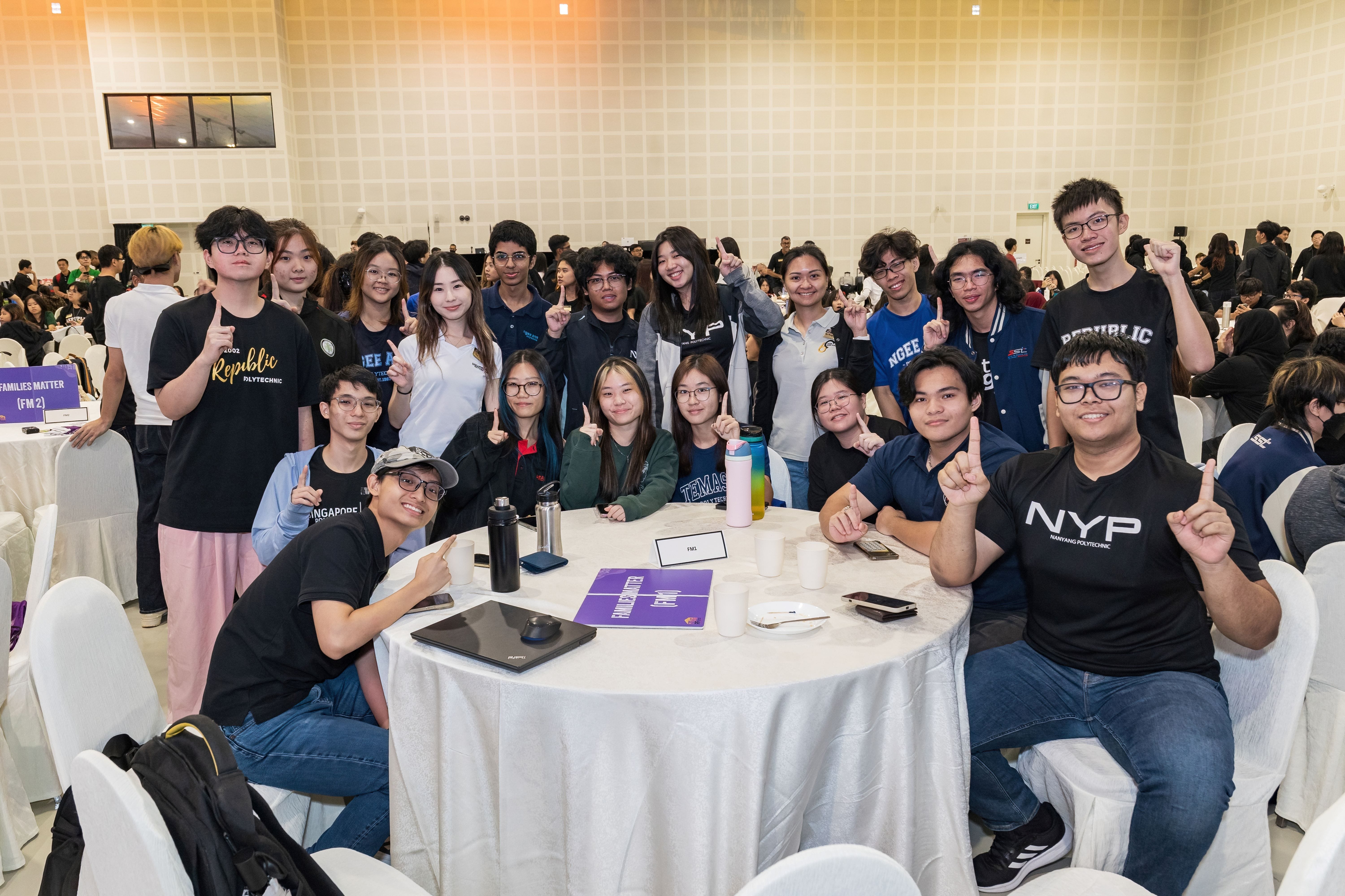 A large group of young students posing and pointing one finger up around a round table at an indoor event.