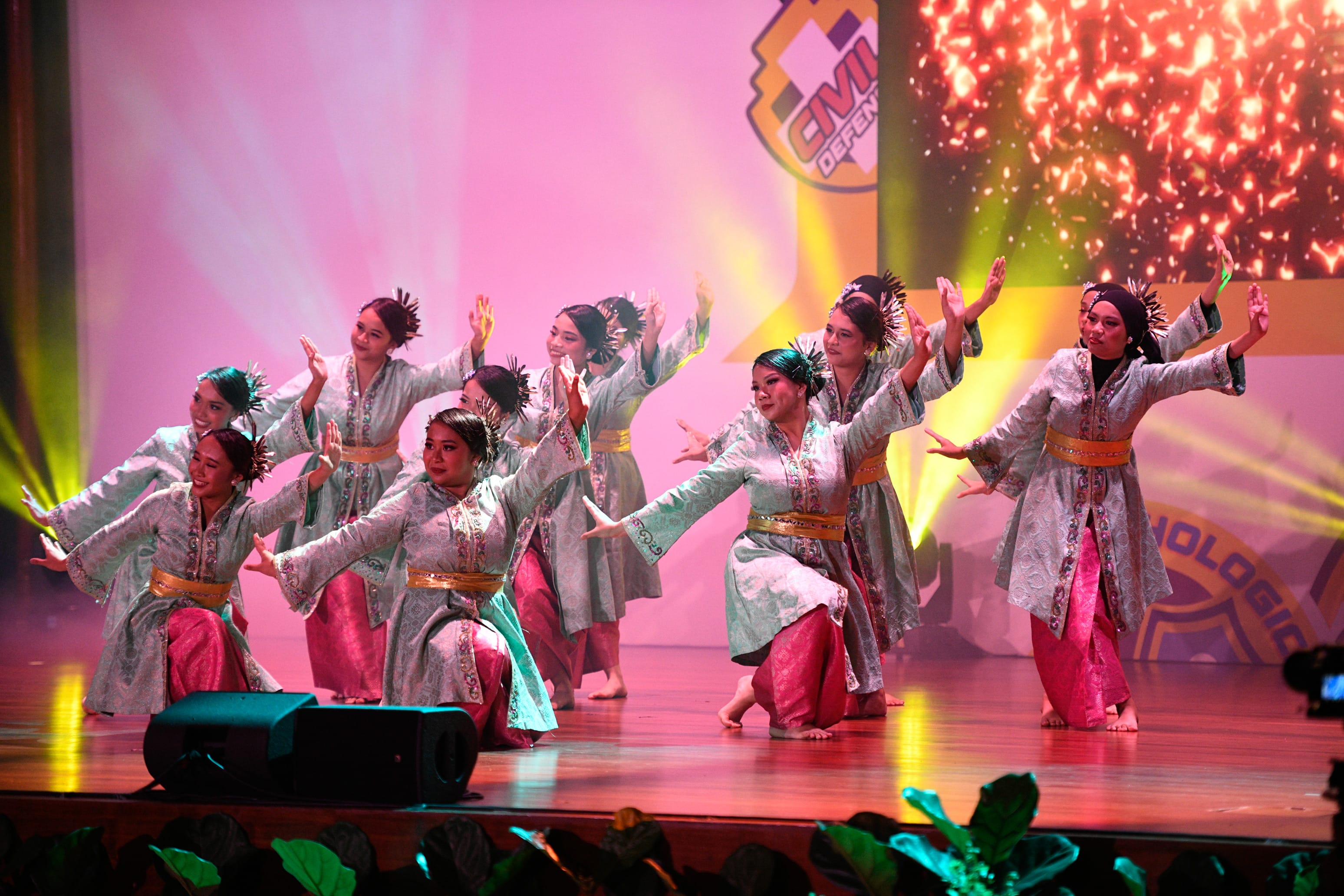 Group of women in silver and pink traditional costumes performing a synchronized dance on a lit stage.