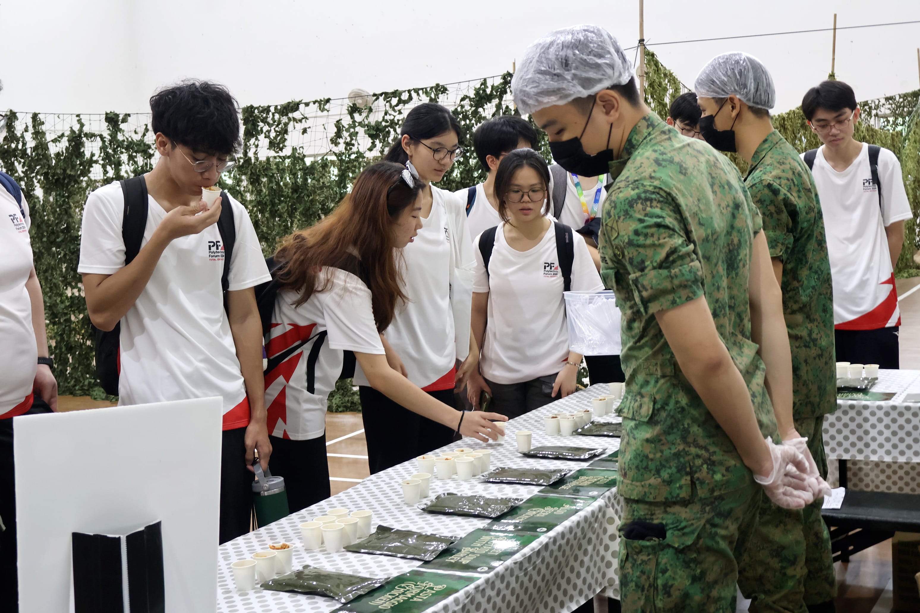 Students in white shirts sample food from cups on a table, guided by a person in military camouflage uniform wearing gloves and hairnet.