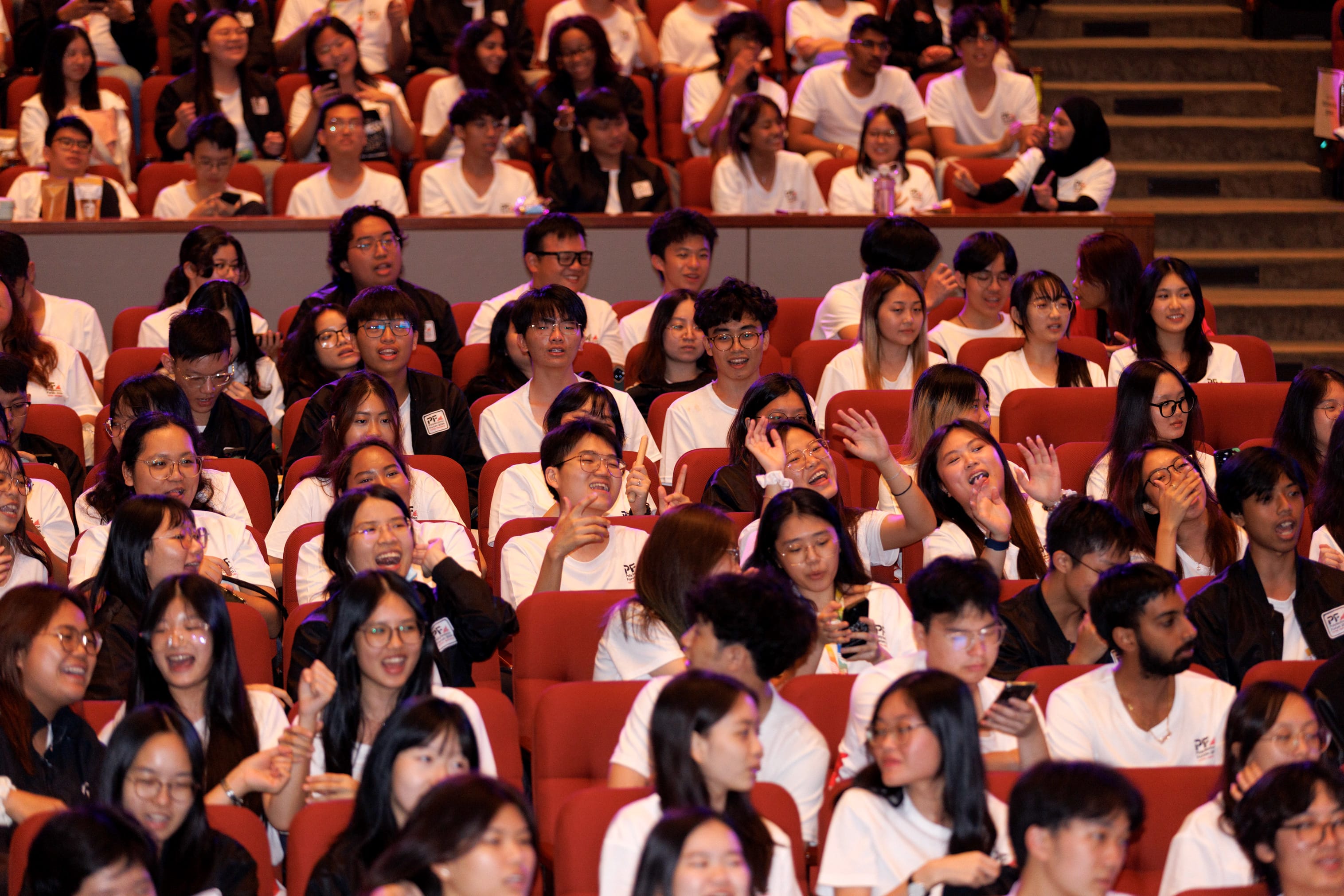 A large crowd of young people in white shirts sit in red auditorium seats, laughing and cheering.