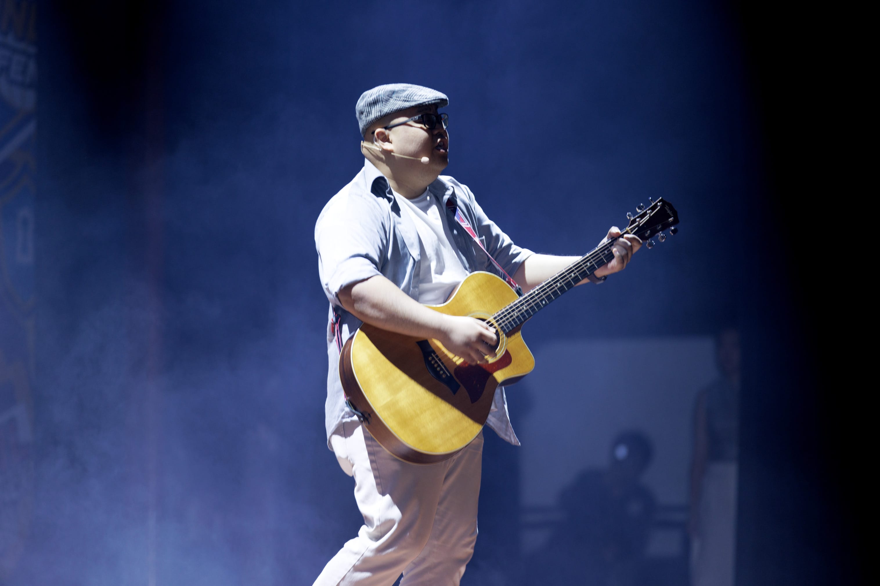 Man in a flat cap and sunglasses playing an acoustic guitar on stage with blue smoky lighting.