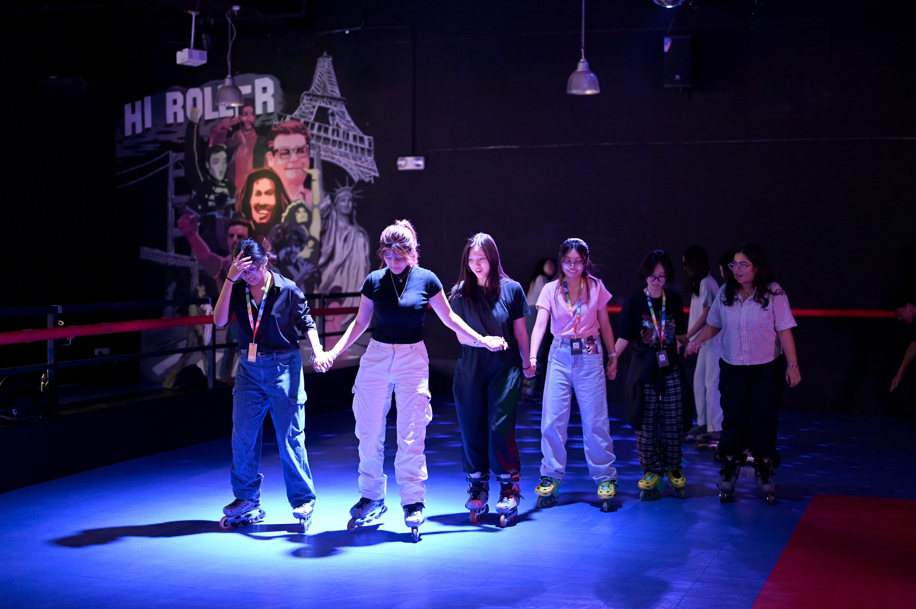A group of young people holding hands while roller skating in a dim rink with a mural on the dark wall behind them.