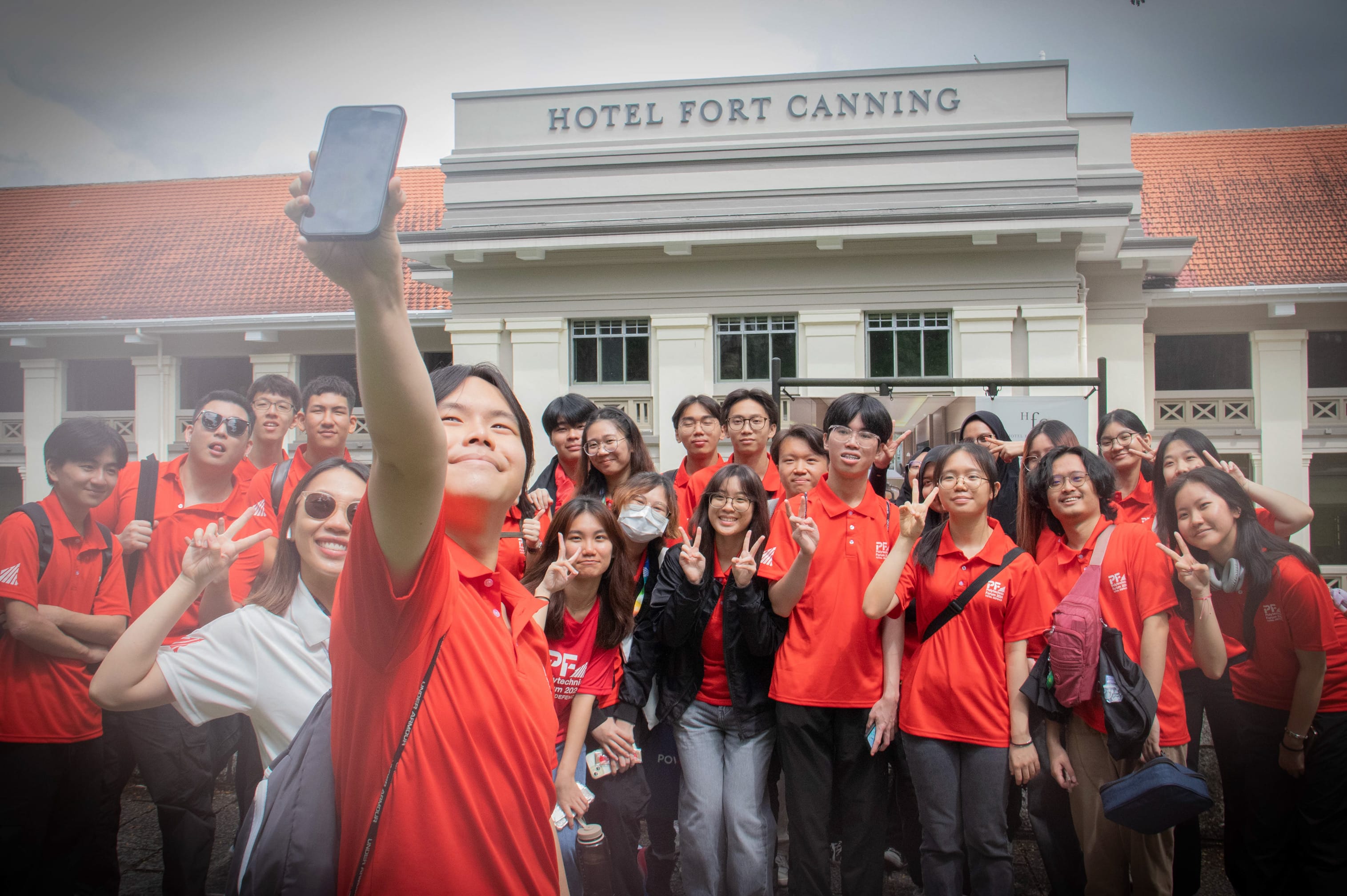 A large group in red polo shirts takes a selfie in front of Hotel Fort Canning, many flashing peace signs.