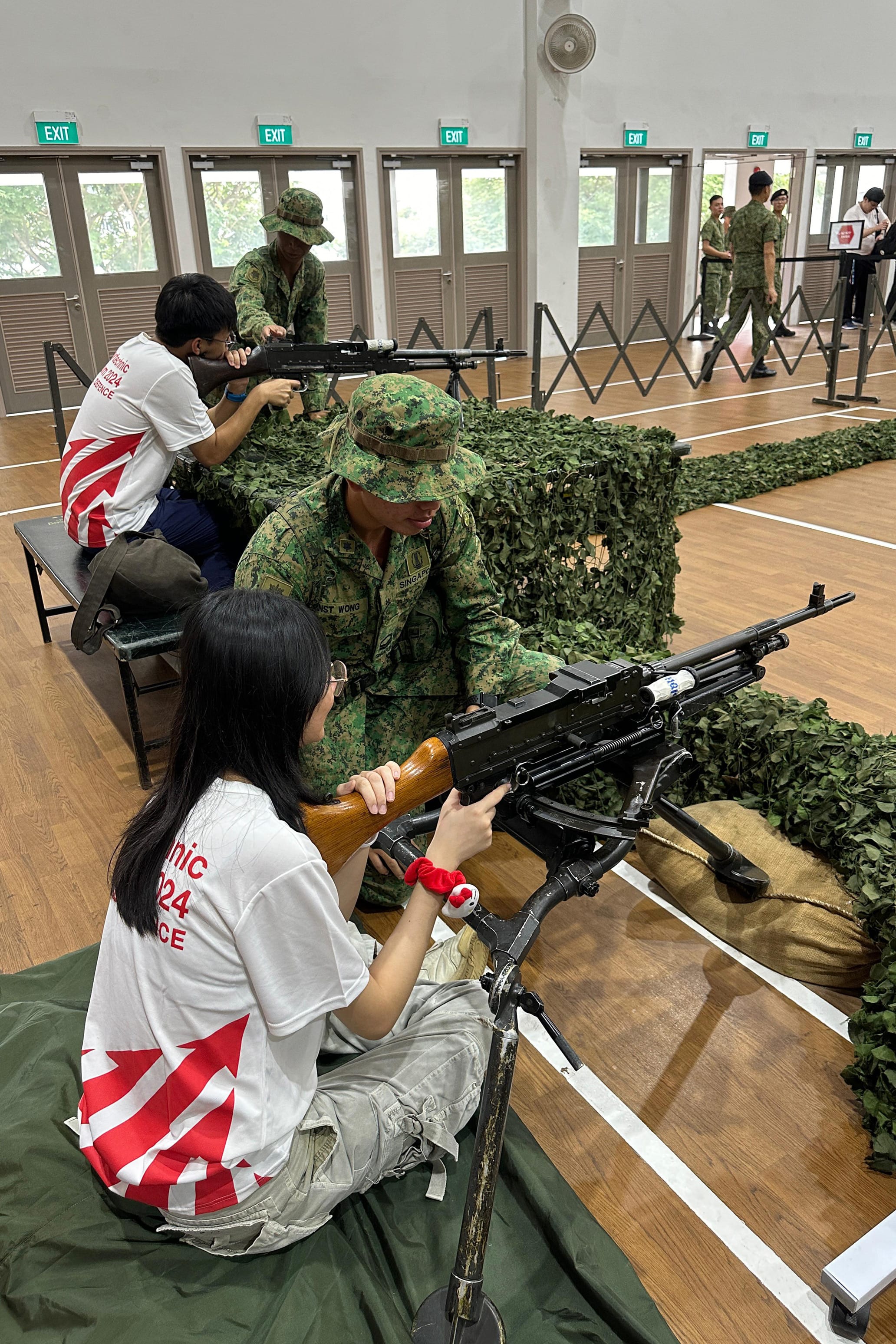 Two civilians handle mounted machine guns while soldiers in camouflage assist them indoors at a military open house display.
