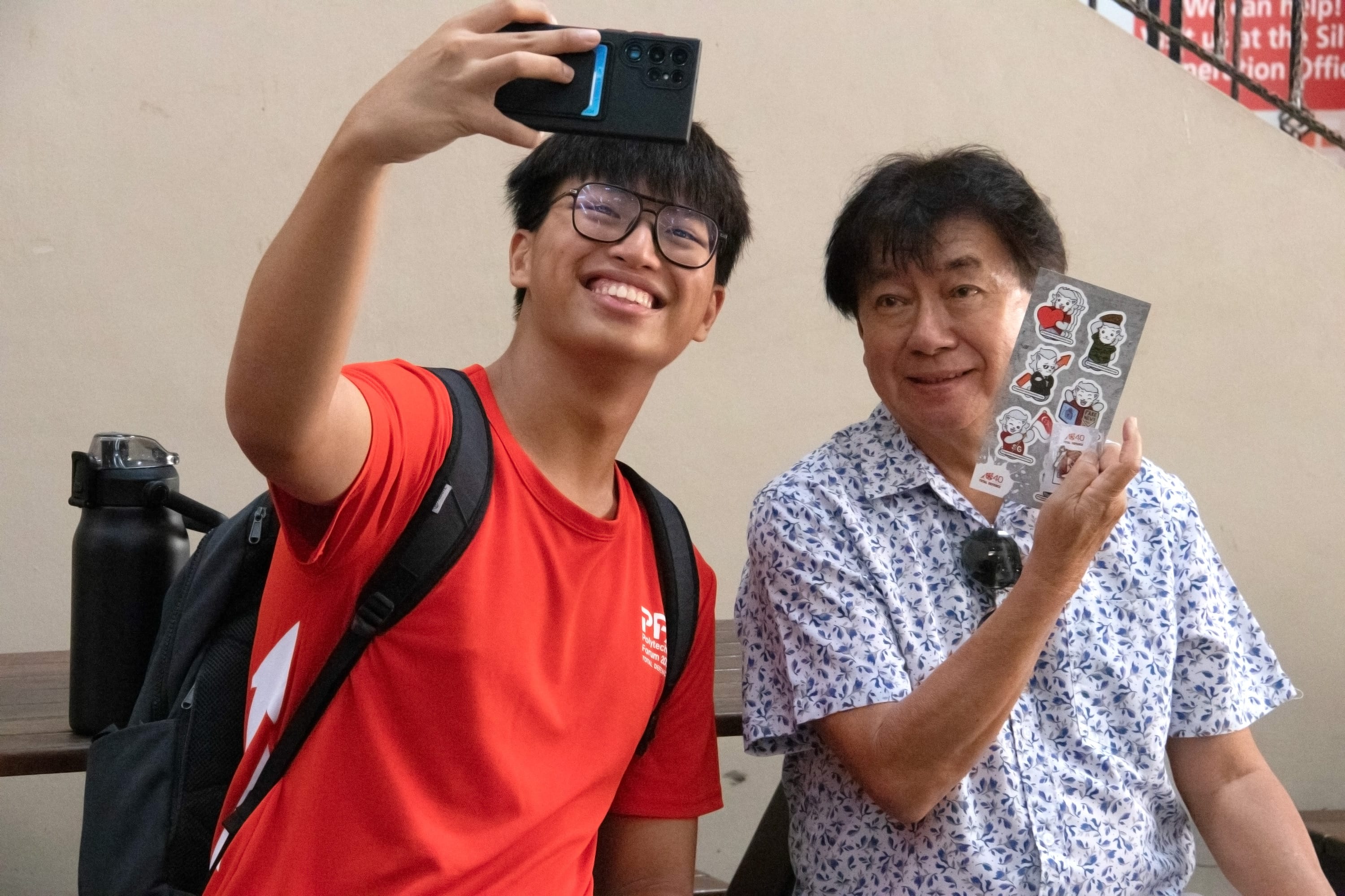 Young man in red shirt takes a selfie with an older man holding a sticker sheet, both smiling indoors.
