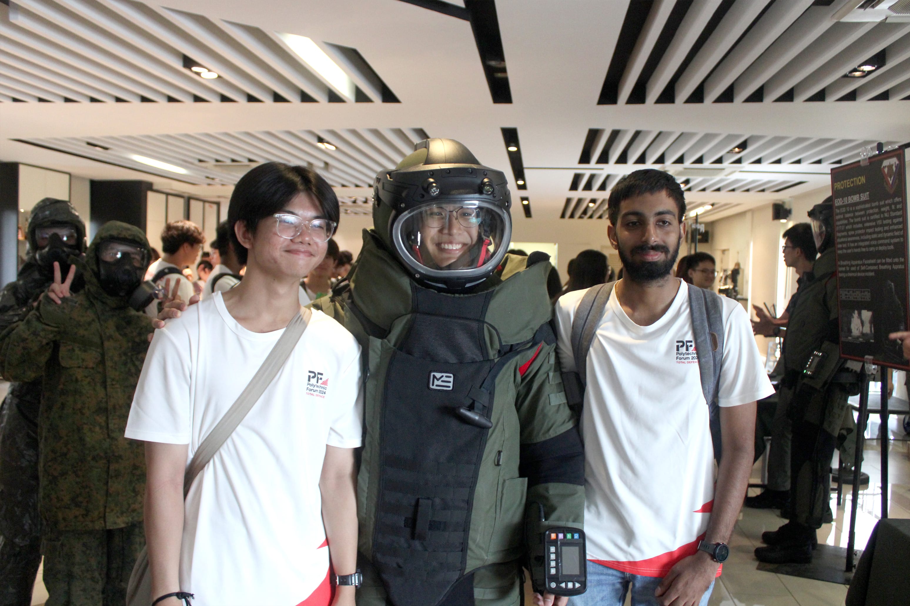 Three people pose together indoors, with the middle person wearing a full bomb disposal suit and helmet.