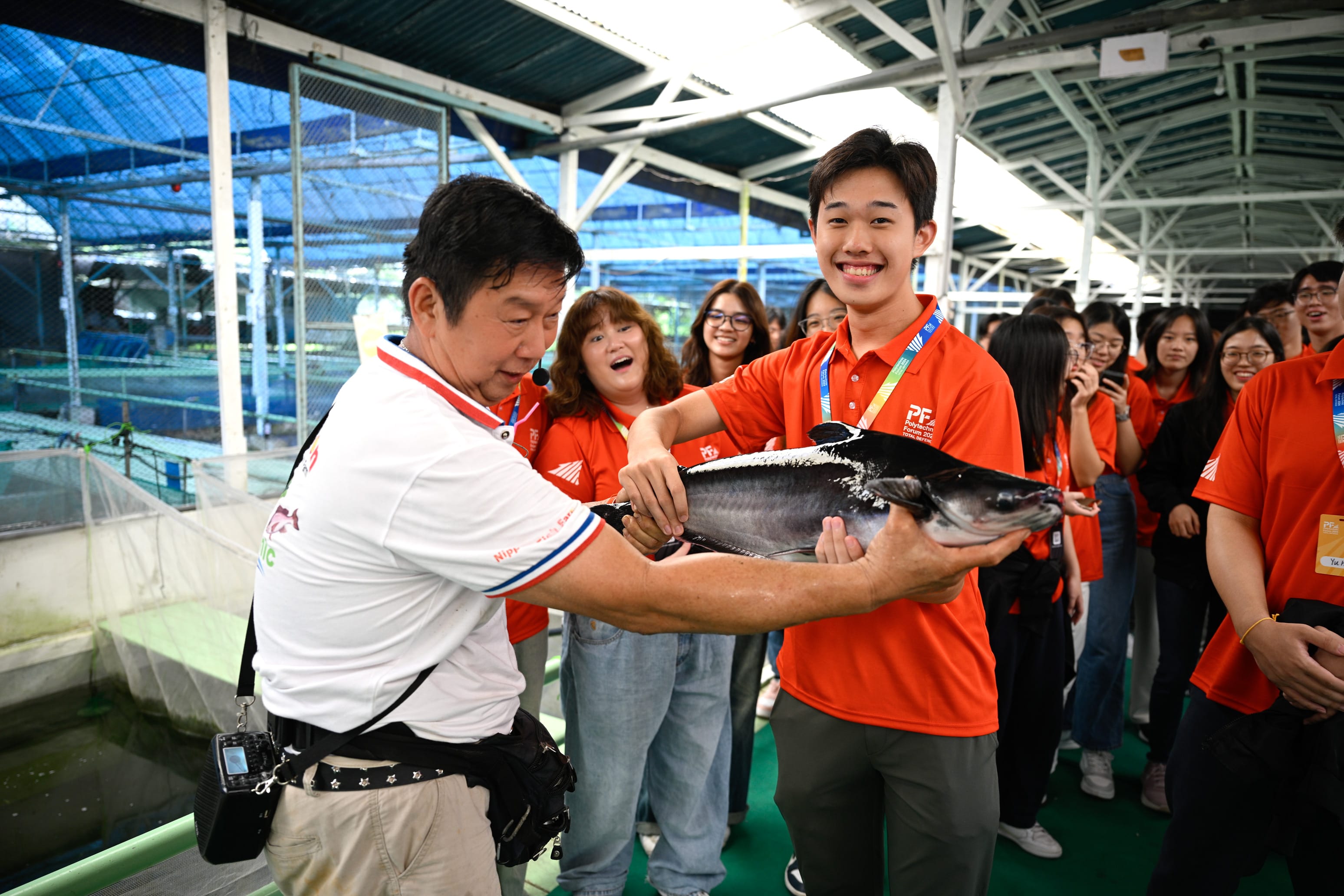Two men hold a large dark fish together at an aquaculture facility as a group of students in orange shirts watches.