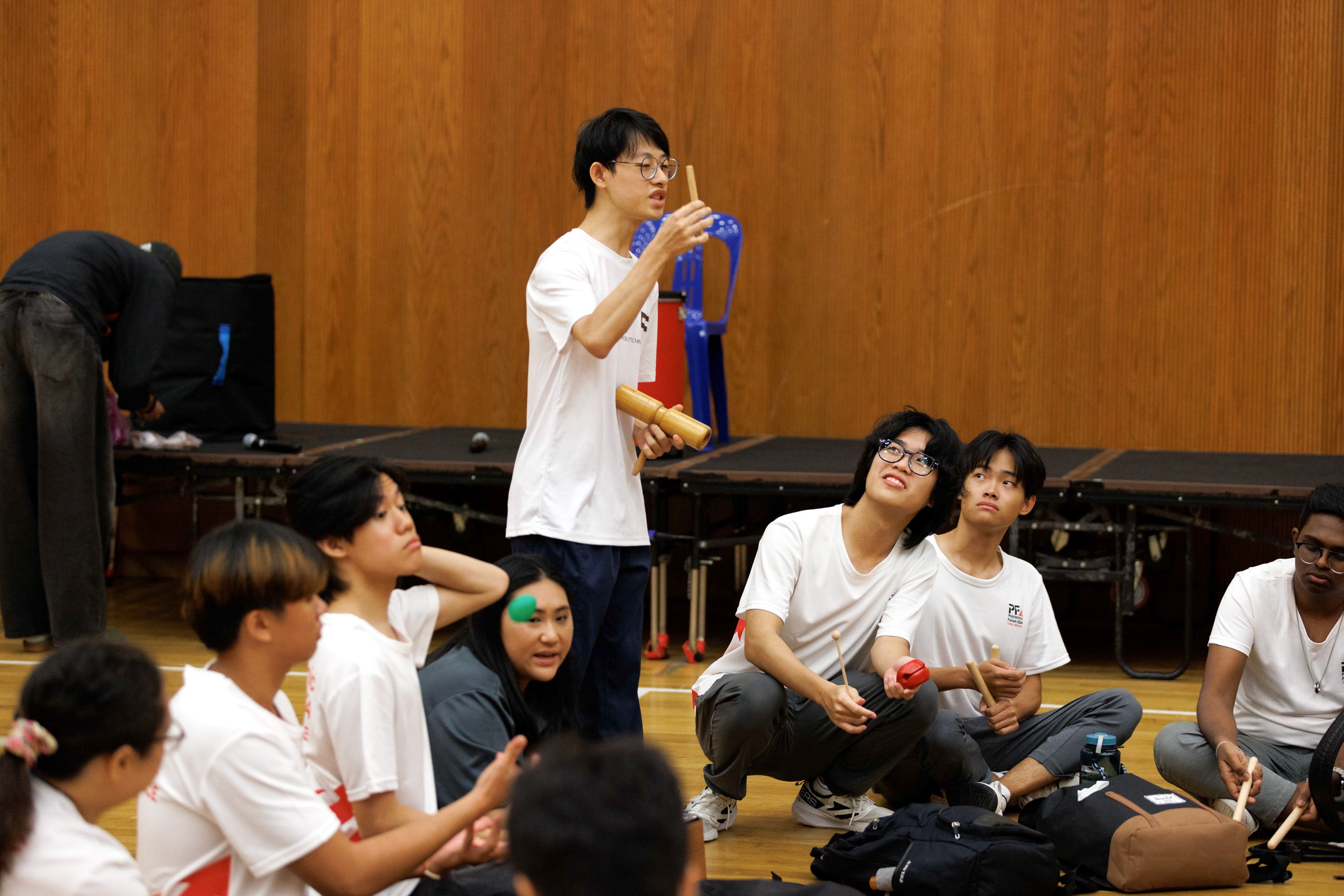 A young man stands holding wooden sticks, demonstrating to a group of students sitting on a gymnasium floor.