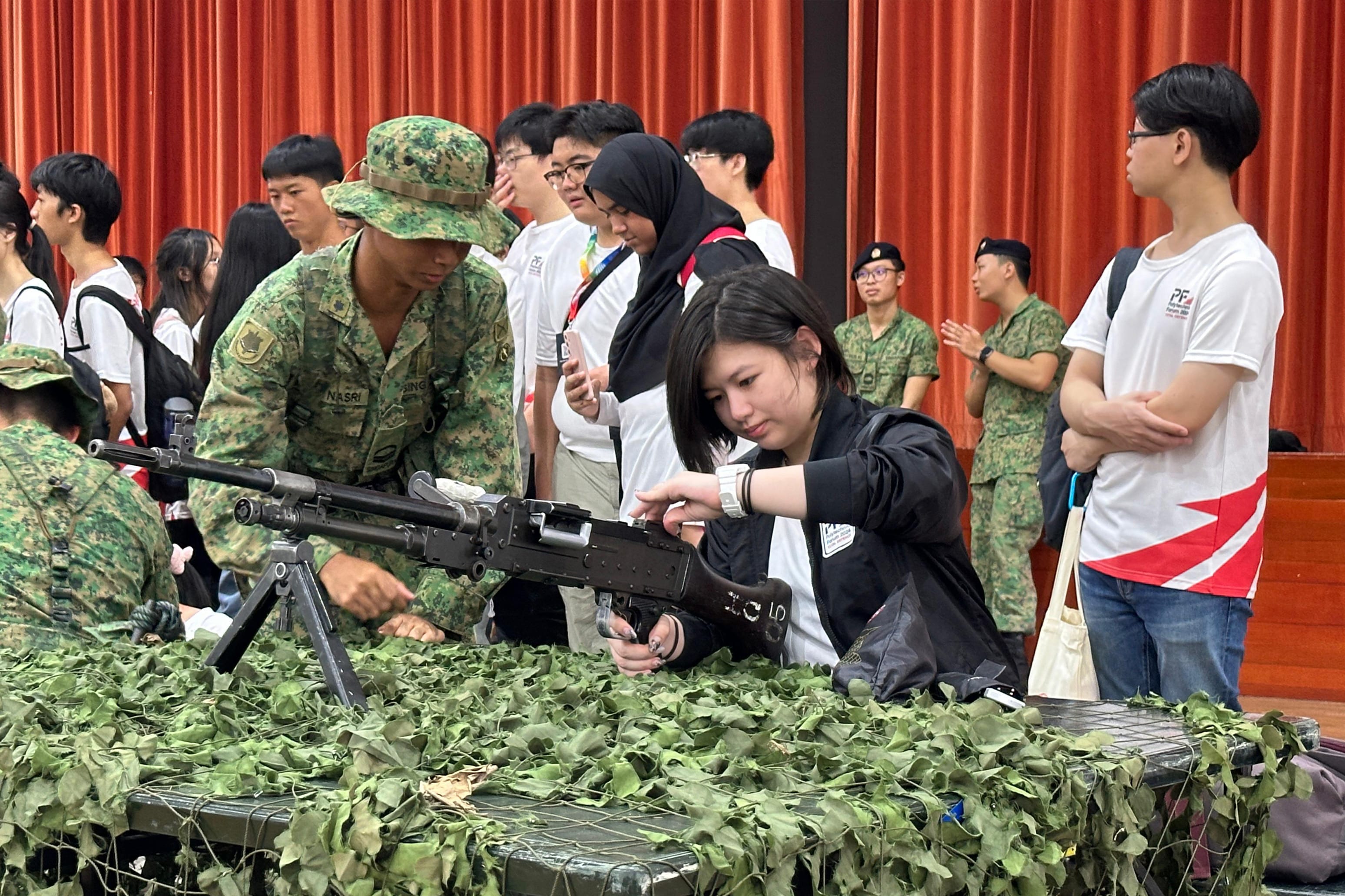 A soldier in camouflage guides a young woman handling a machine gun on a camouflage-covered table at a military exhibition.
