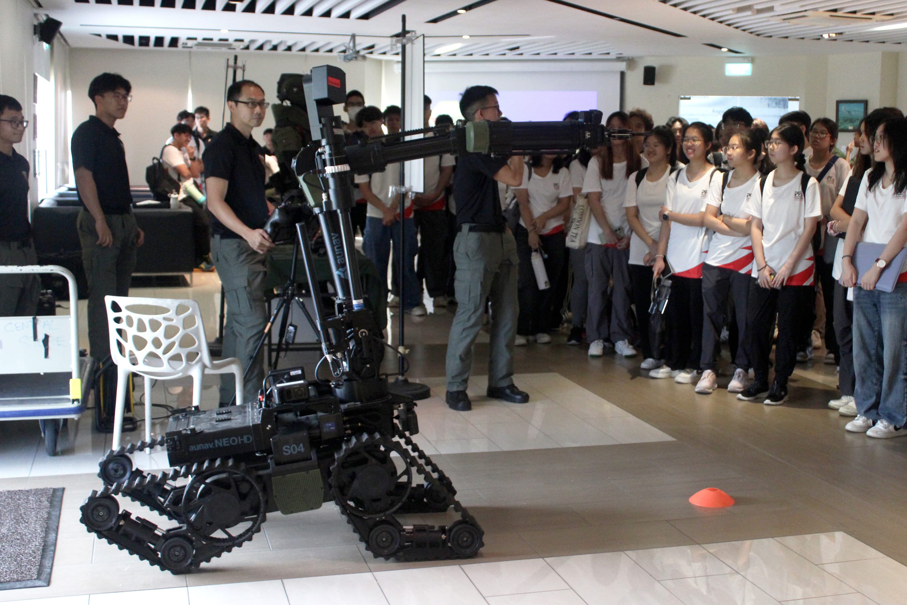 A tracked robot with a robotic arm is demonstrated to a large group of students watching in an indoor hall.
