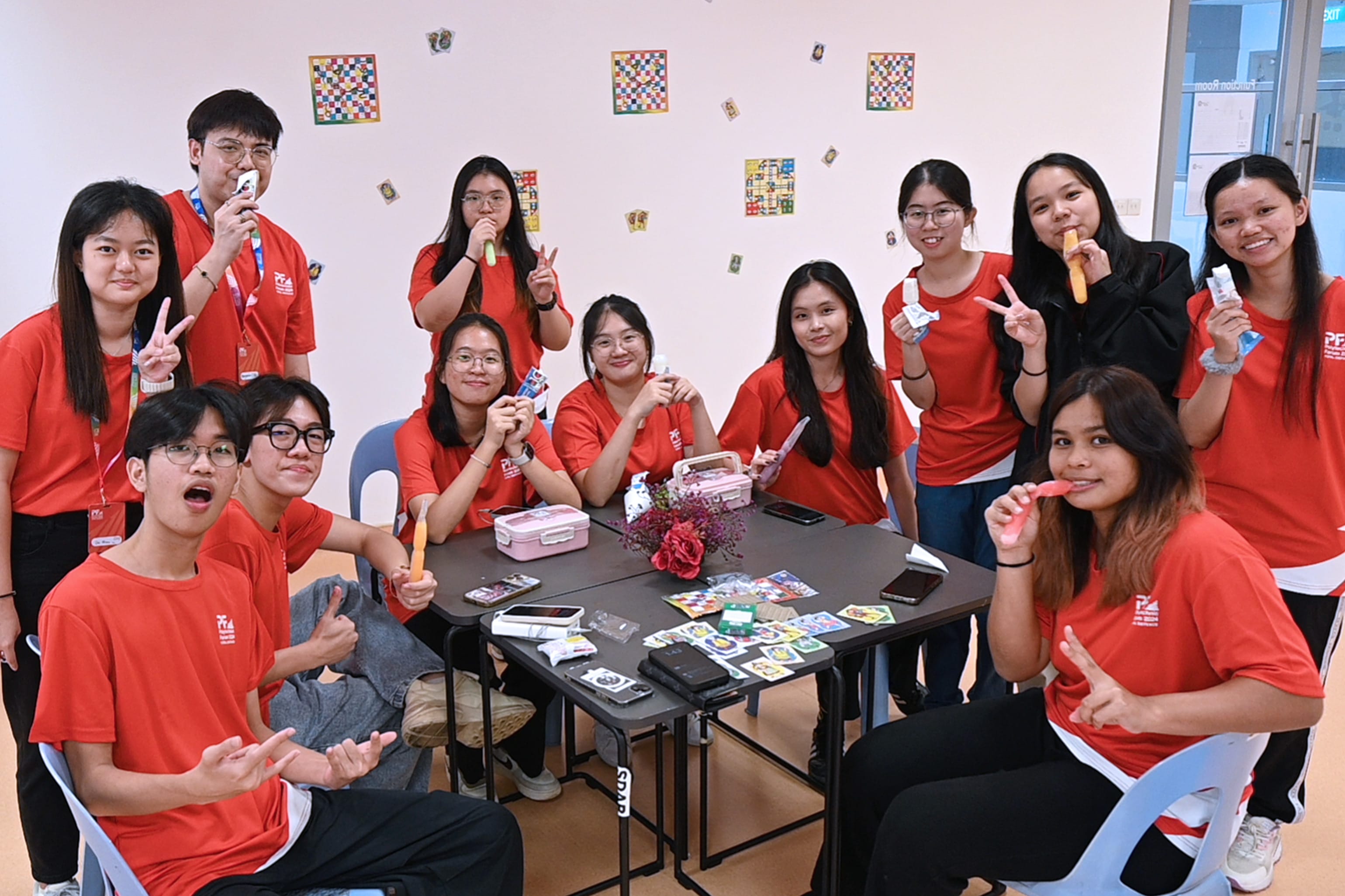 A group of young people in red shirts pose and smile around a table with cards and snacks in a decorated room.