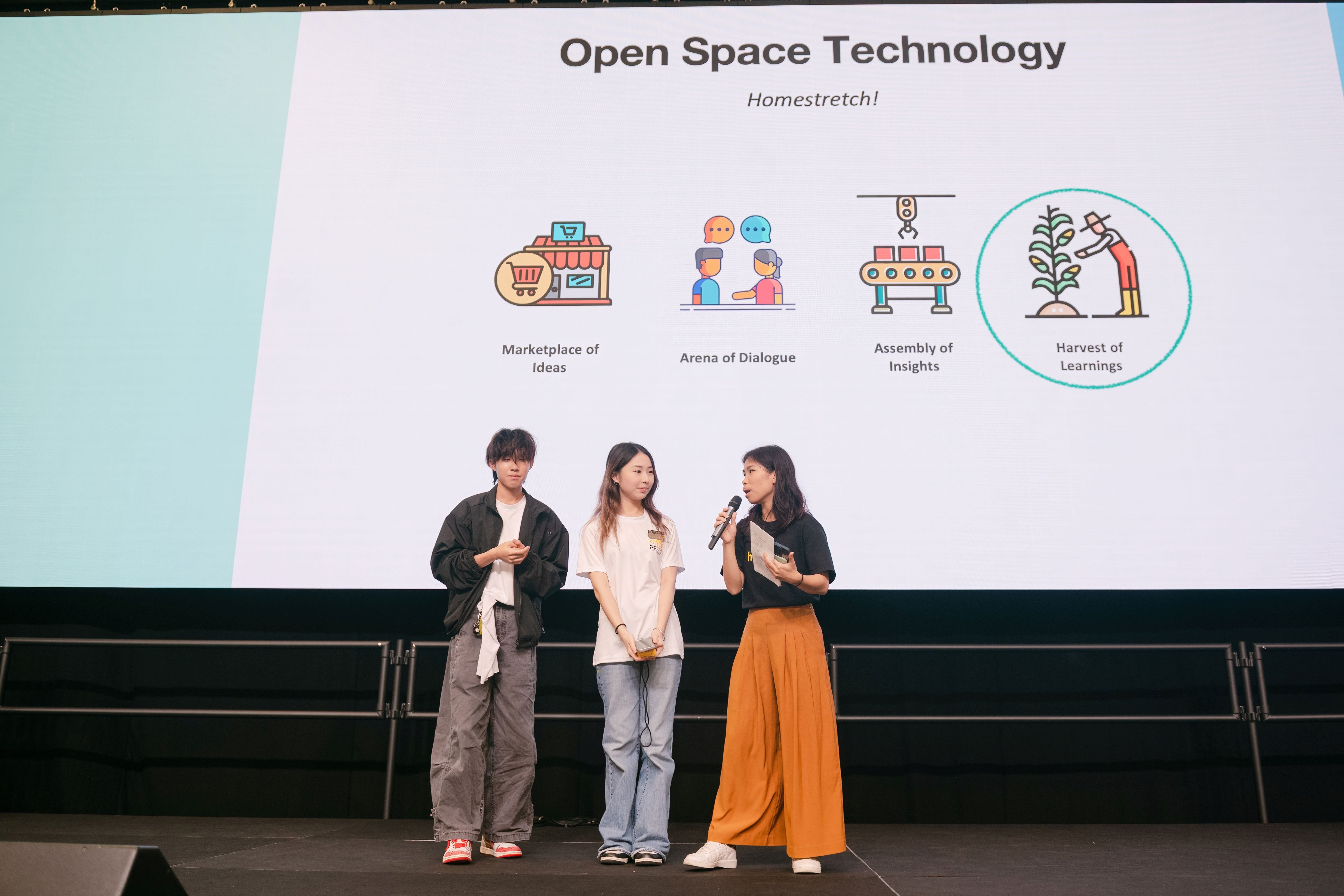 Three people stand on stage before a slide titled Open Space Technology showing four icons and concepts.