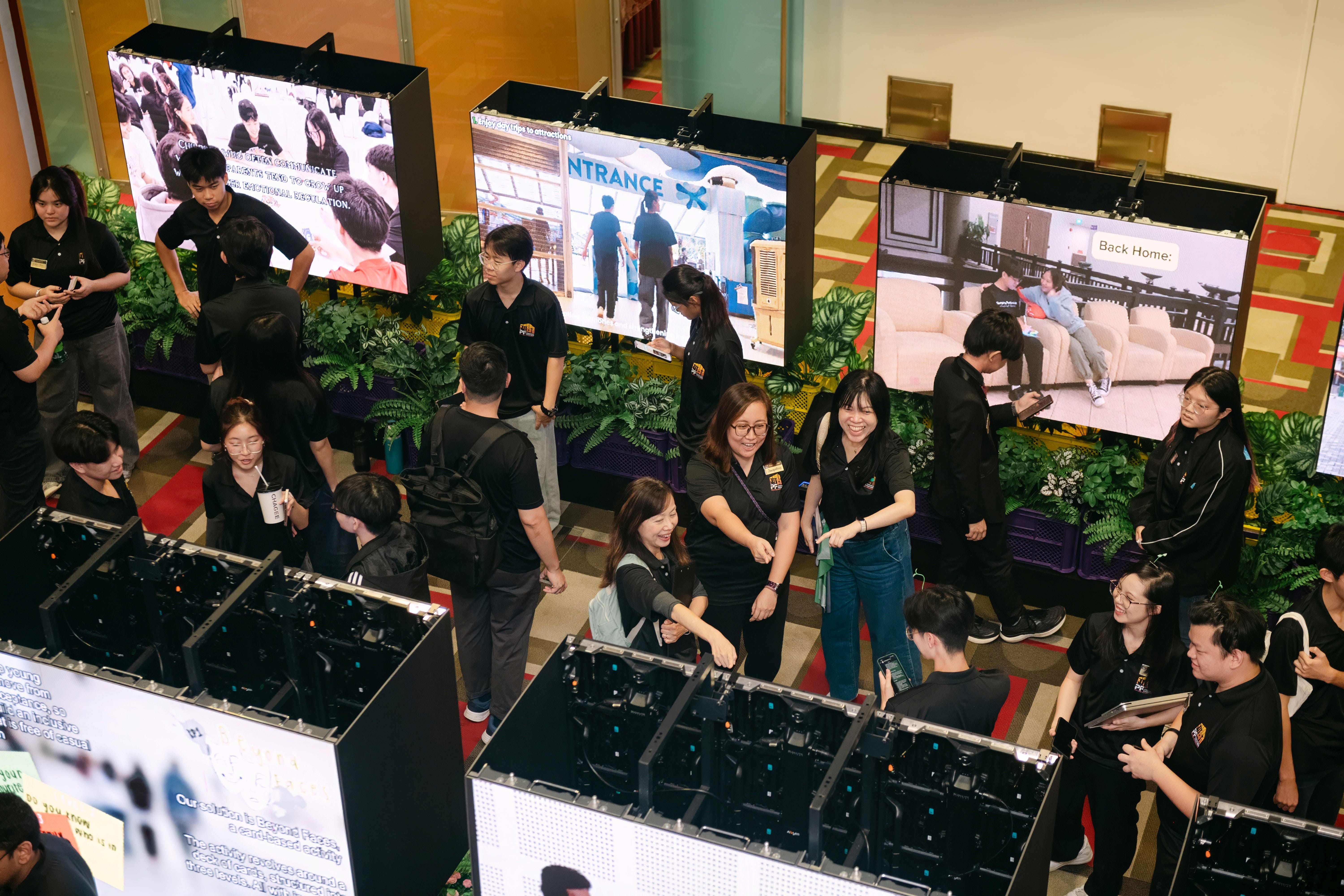 Group of young people in black uniforms gathered around large display screens and equipment at an indoor exhibition.