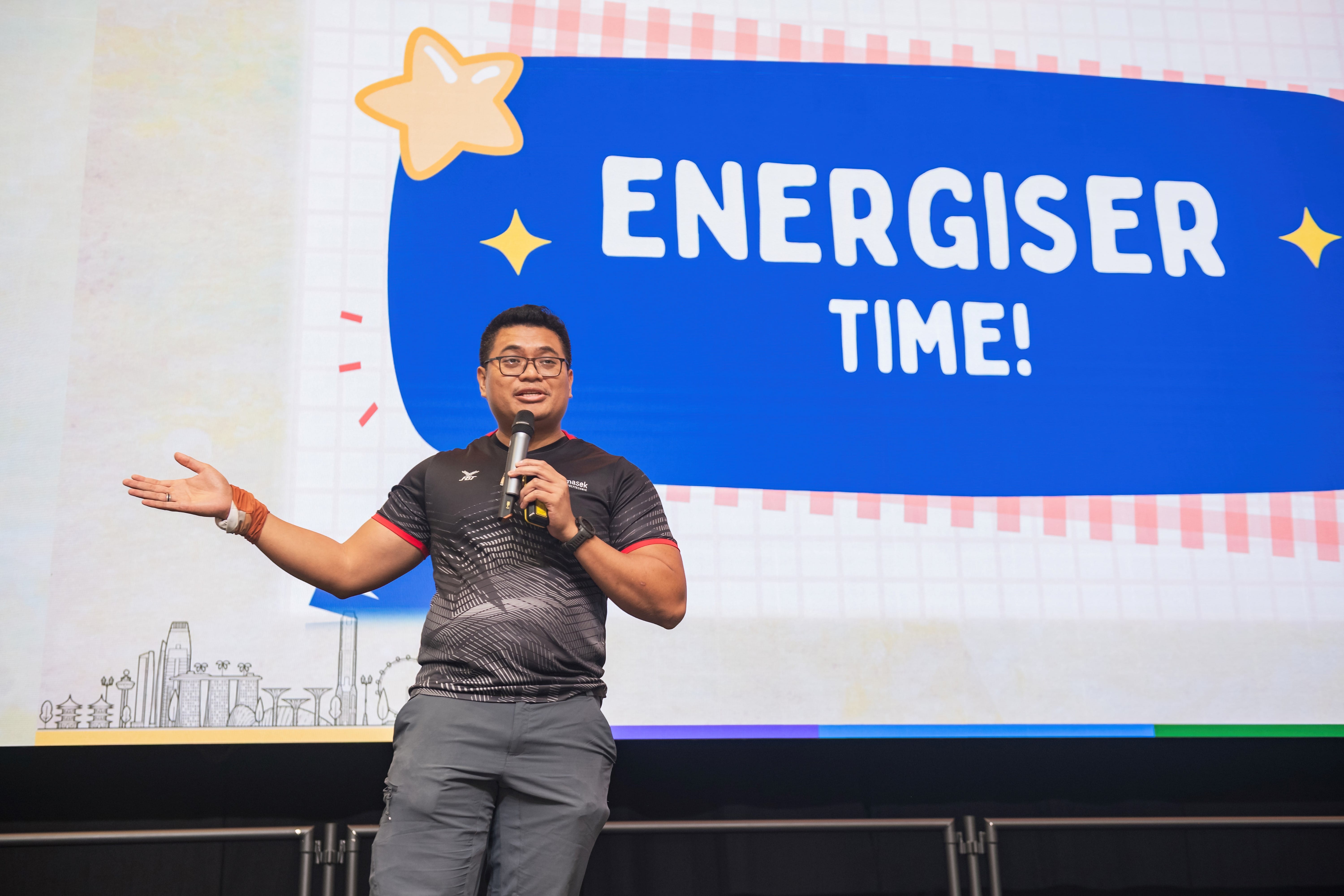 Man with glasses holds a microphone on stage in front of a blue screen reading "ENERGISER TIME!"
