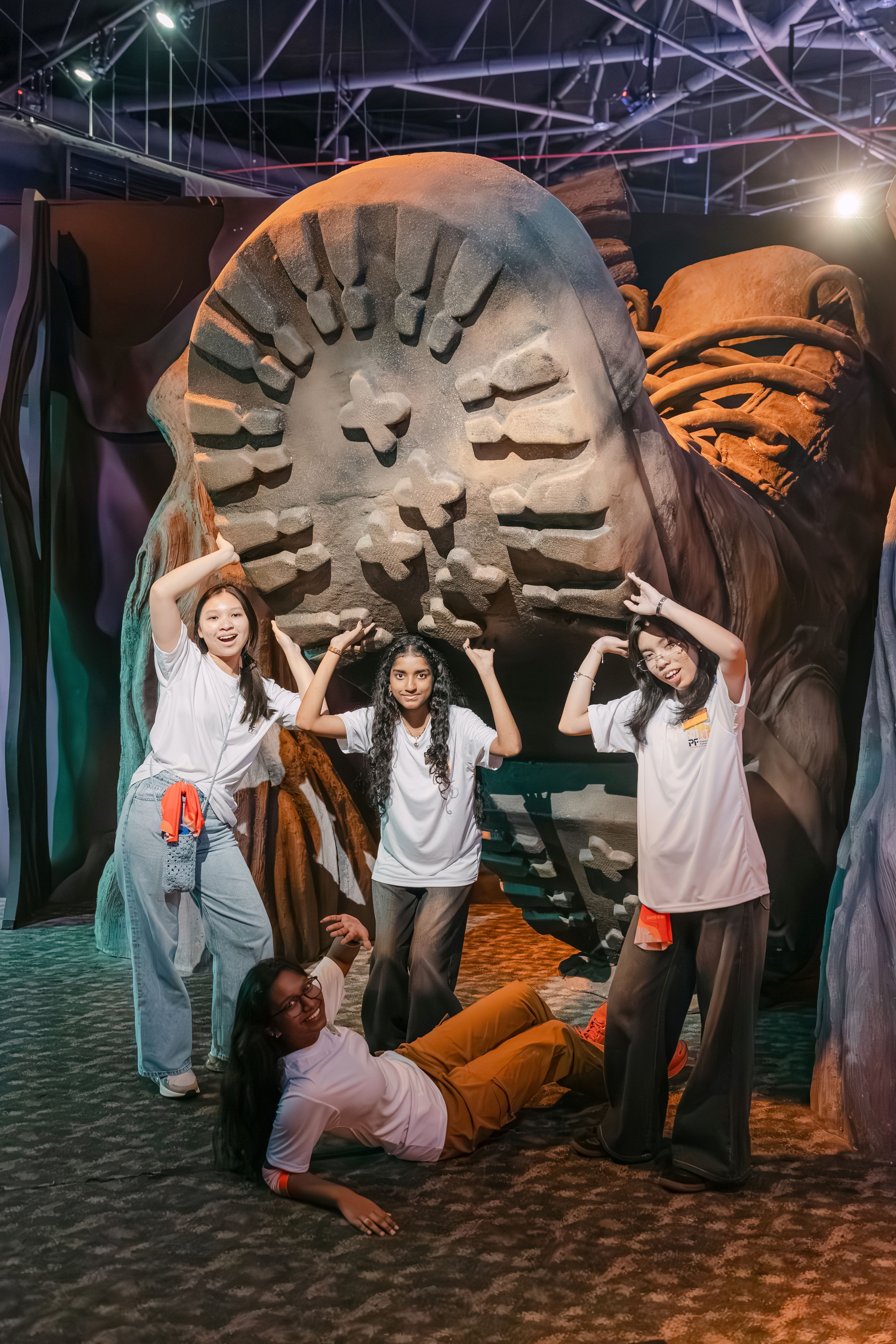 Four young women in white shirts pose playfully in front of a giant oversized boot sole prop indoors.