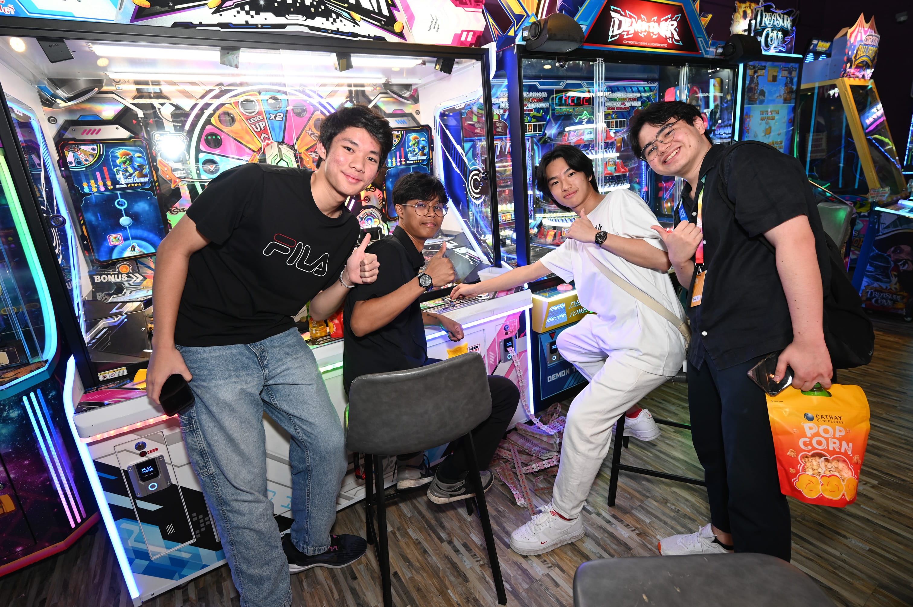 Four young men smile and give thumbs up in a colorful arcade, one holding a Cathay Cineplexes popcorn bucket.
