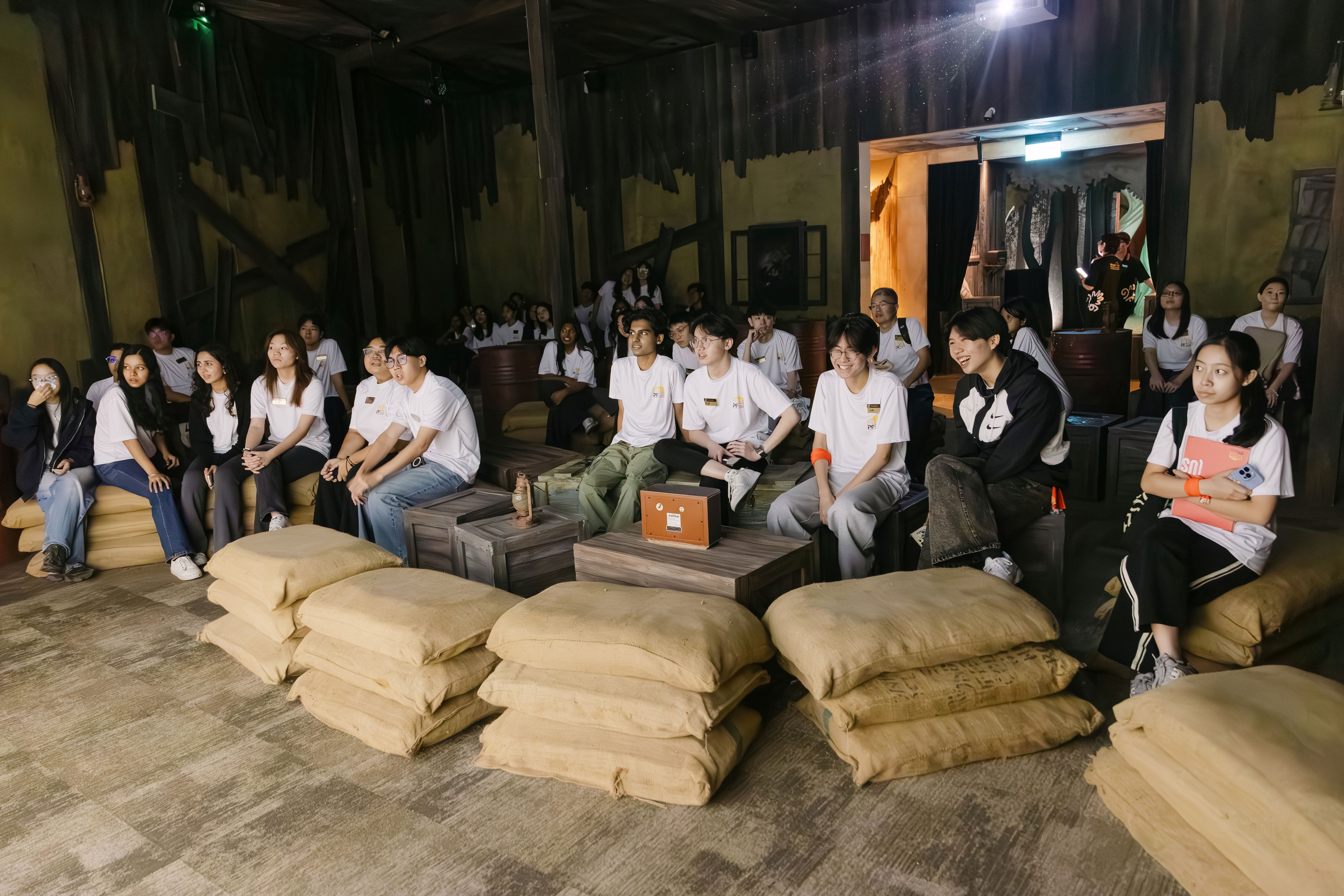 A large group of young people in white t-shirts sit on burlap sacks in a dimly lit rustic themed room.
