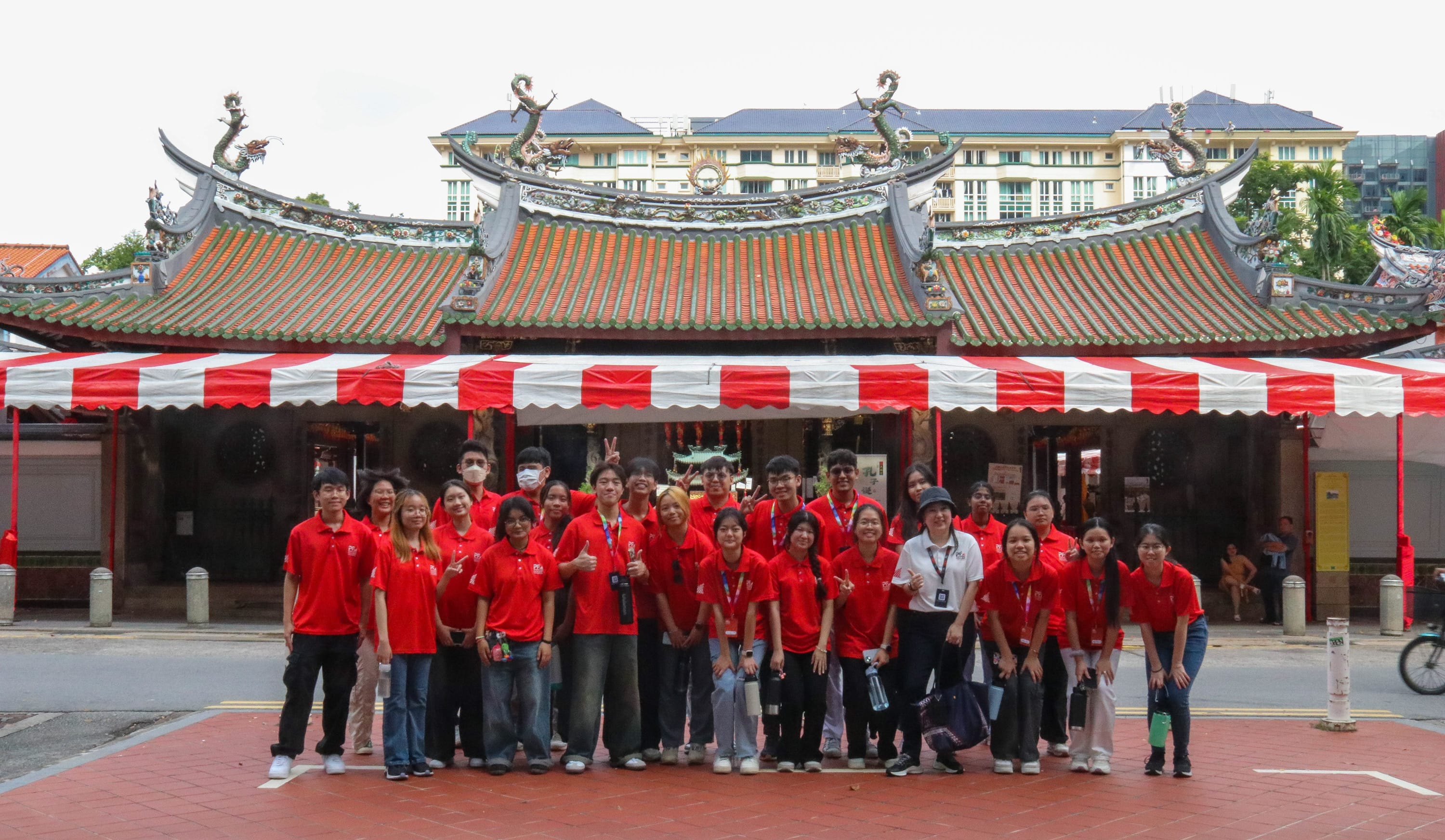 A group of young people mostly in red shirts posing in front of a Chinese temple with dragon roof decorations.