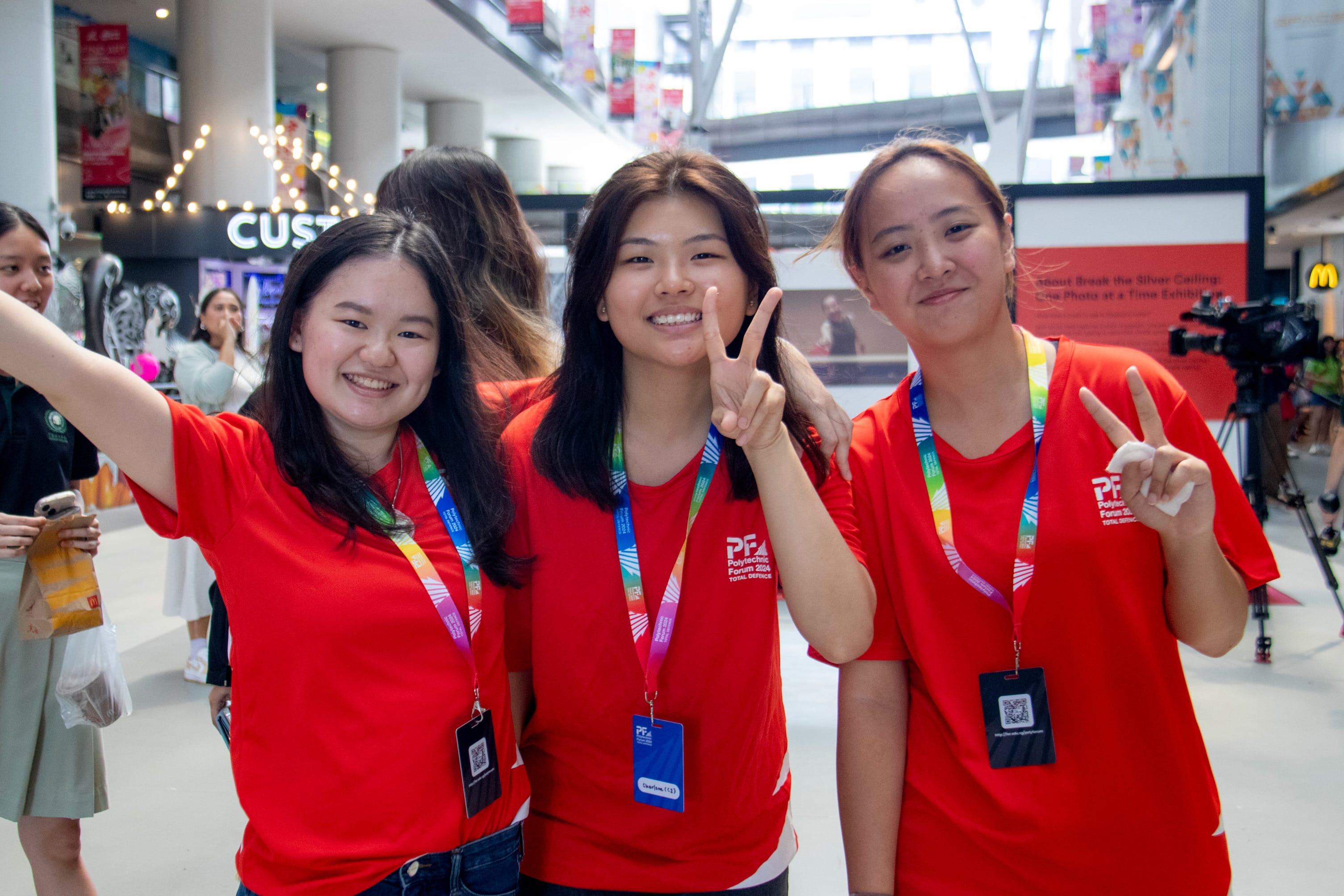 Three young women in red Polytechnic Forum shirts and lanyards smile and flash peace signs indoors.