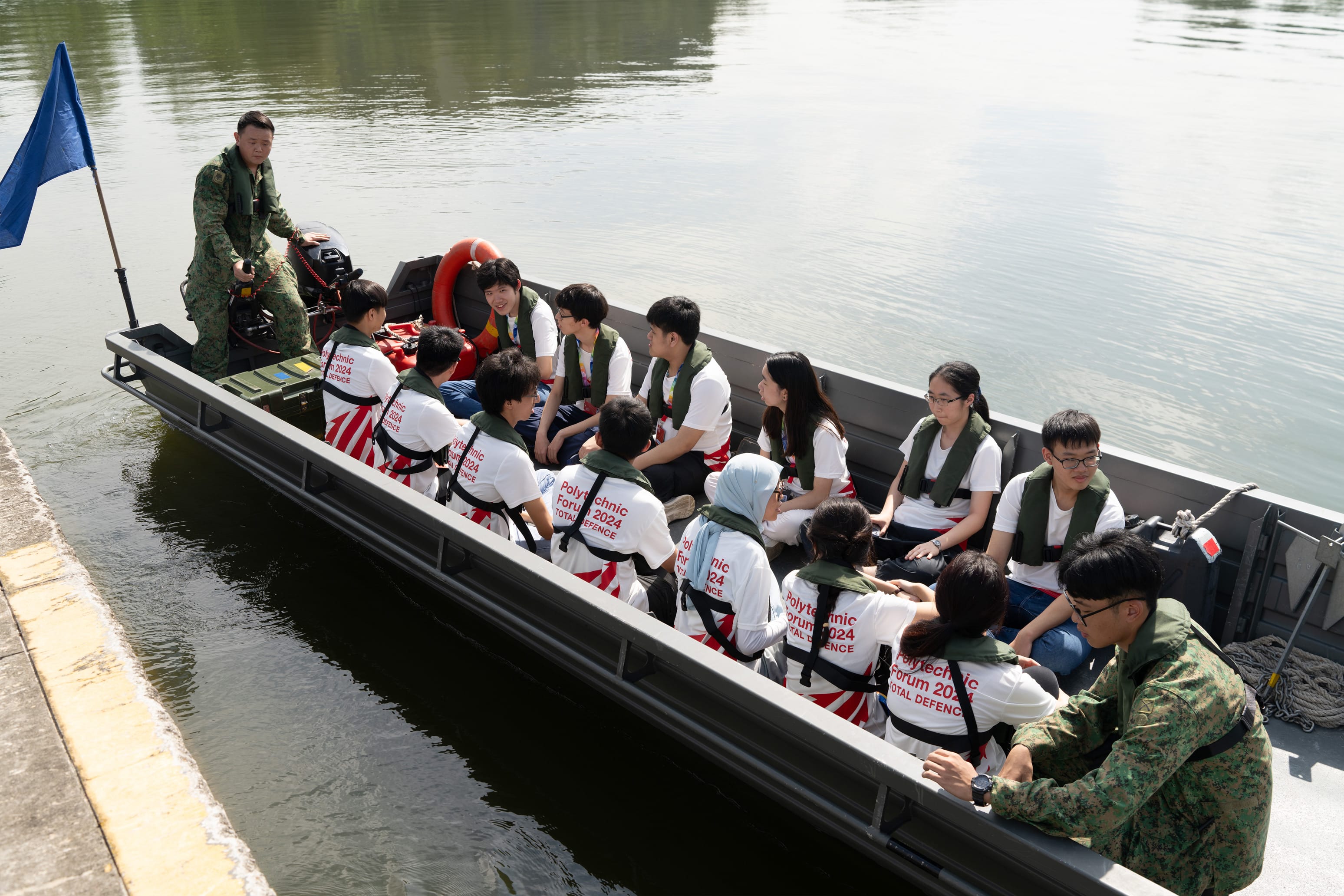 Students in Polytechnic Forum 2024 shirts and life vests sit in a military boat with two soldiers on water.