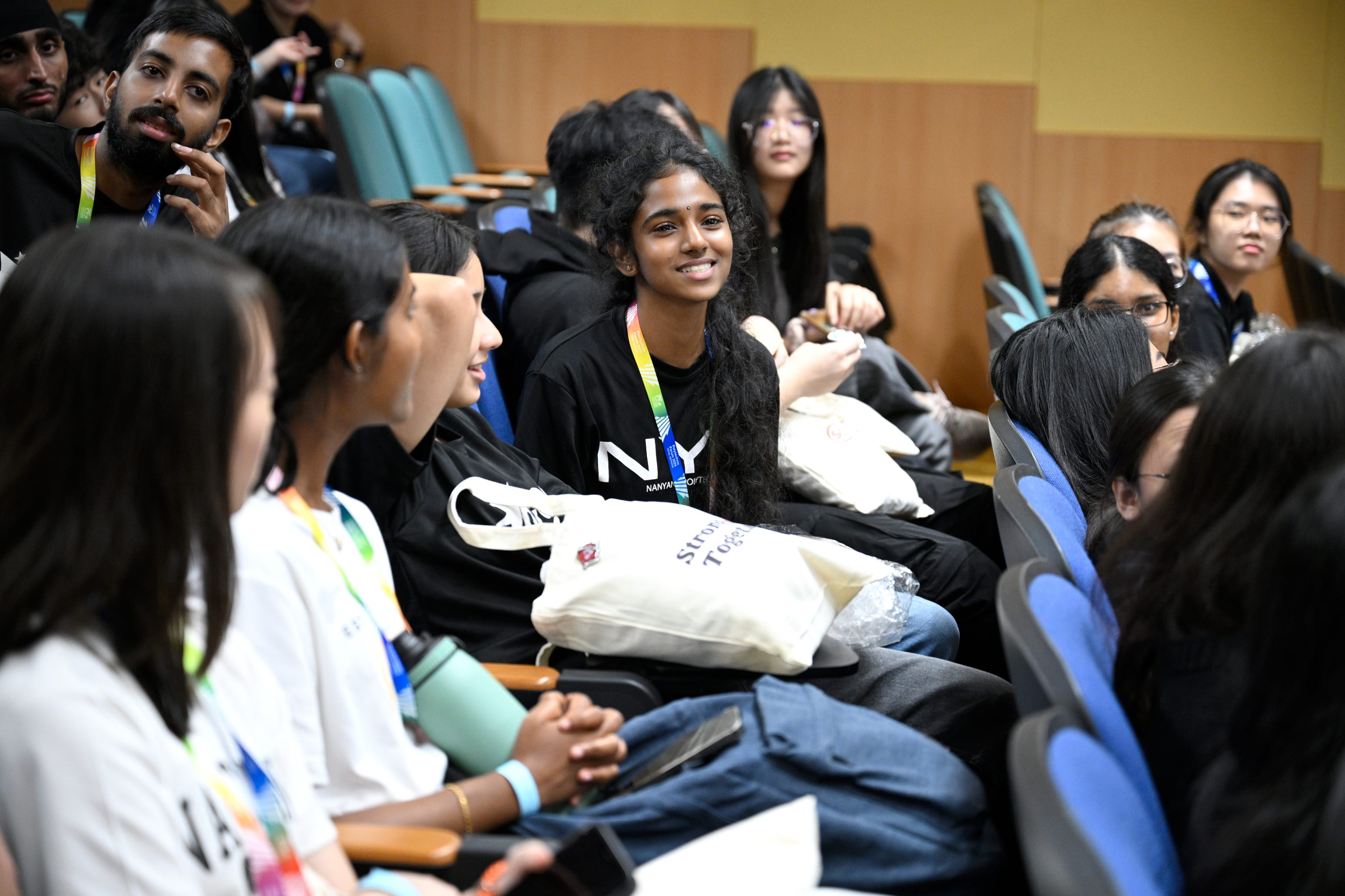 A smiling young woman with a colorful lanyard sits among diverse students in an auditorium, holding a tote bag.