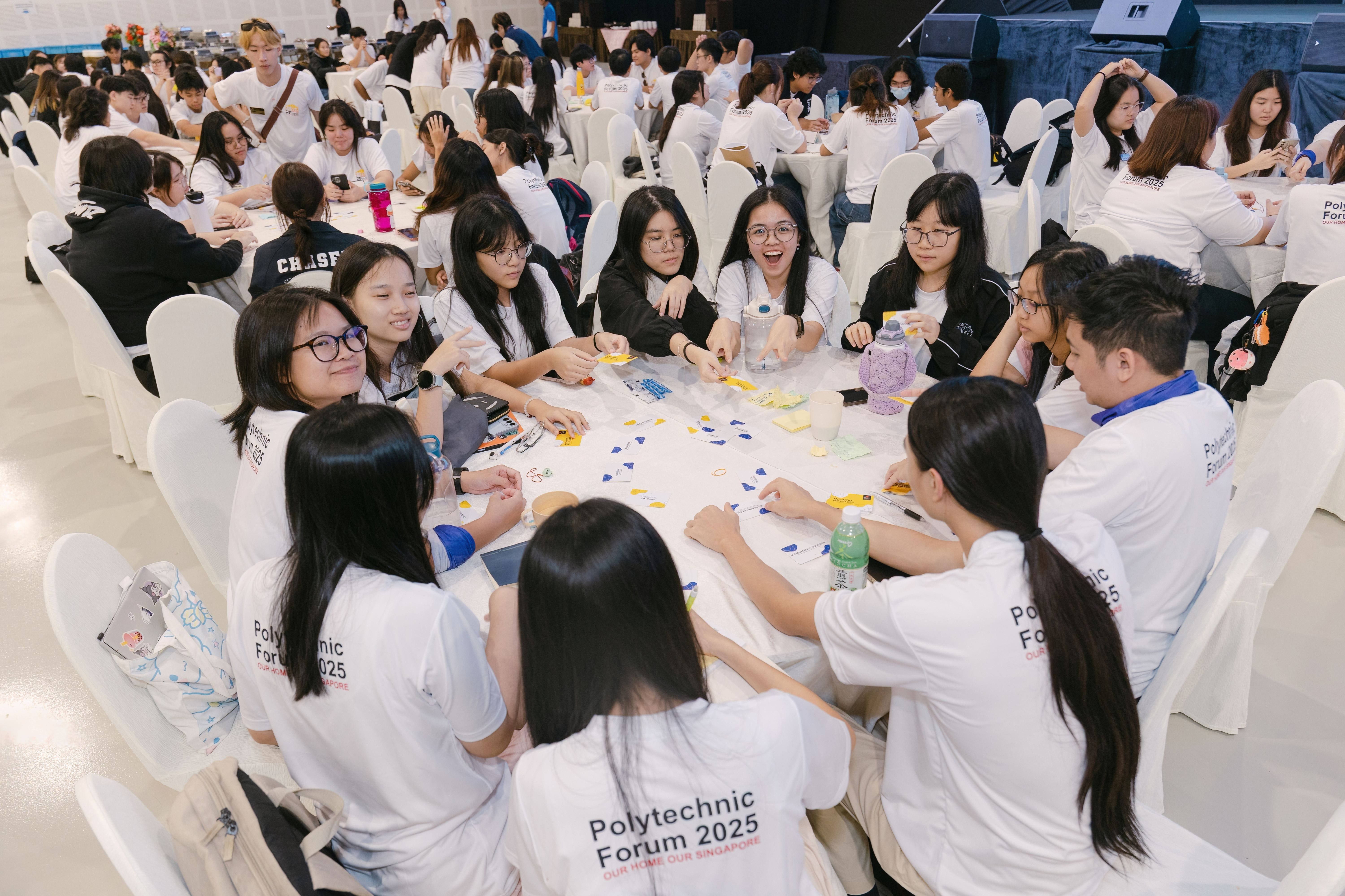 Students in white Polytechnic Forum 2025 shirts sit at round tables doing a group activity in a large hall.