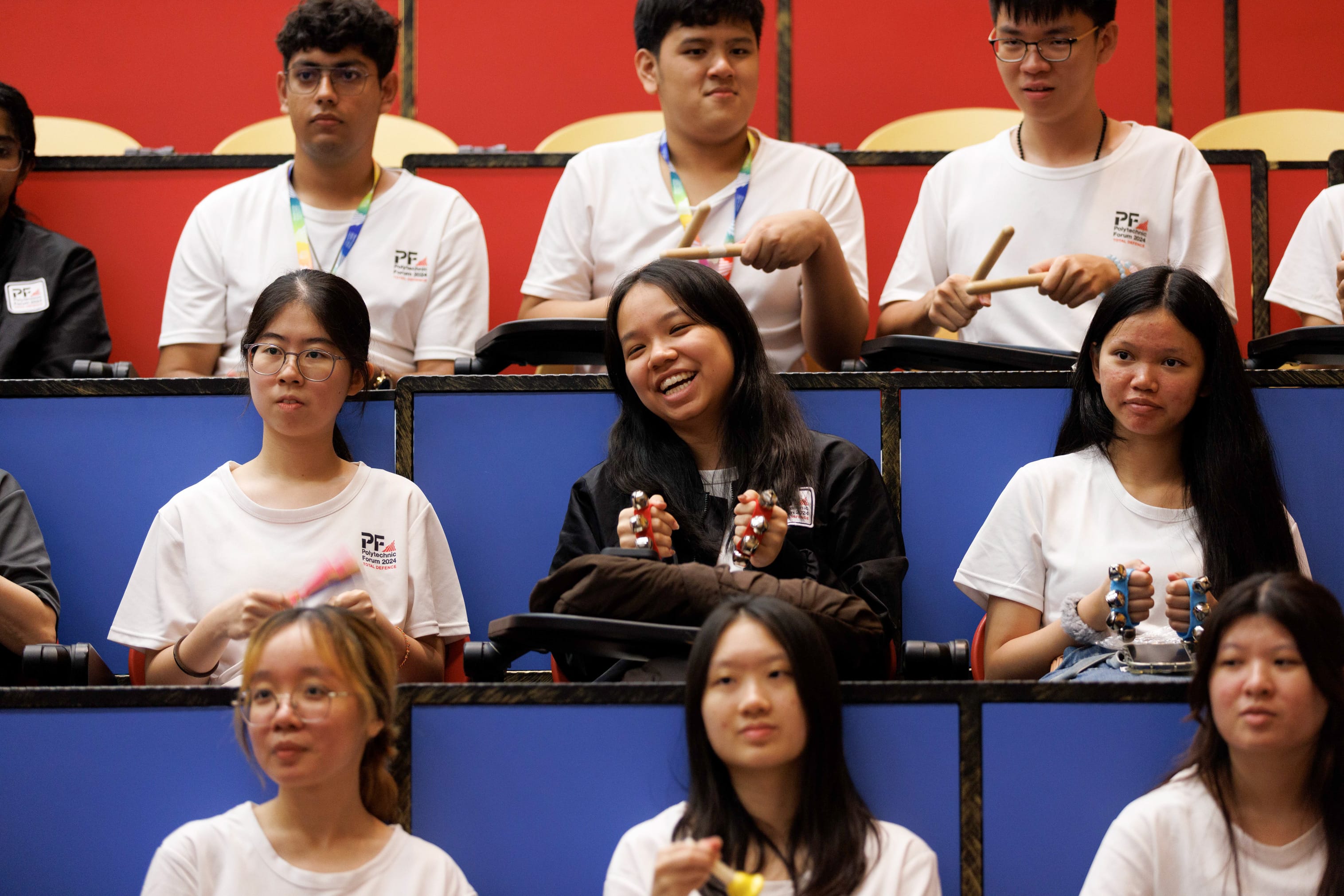 Students in white Polytechnic Forum shirts sit in tiered seats, some smiling and holding small percussion instruments.