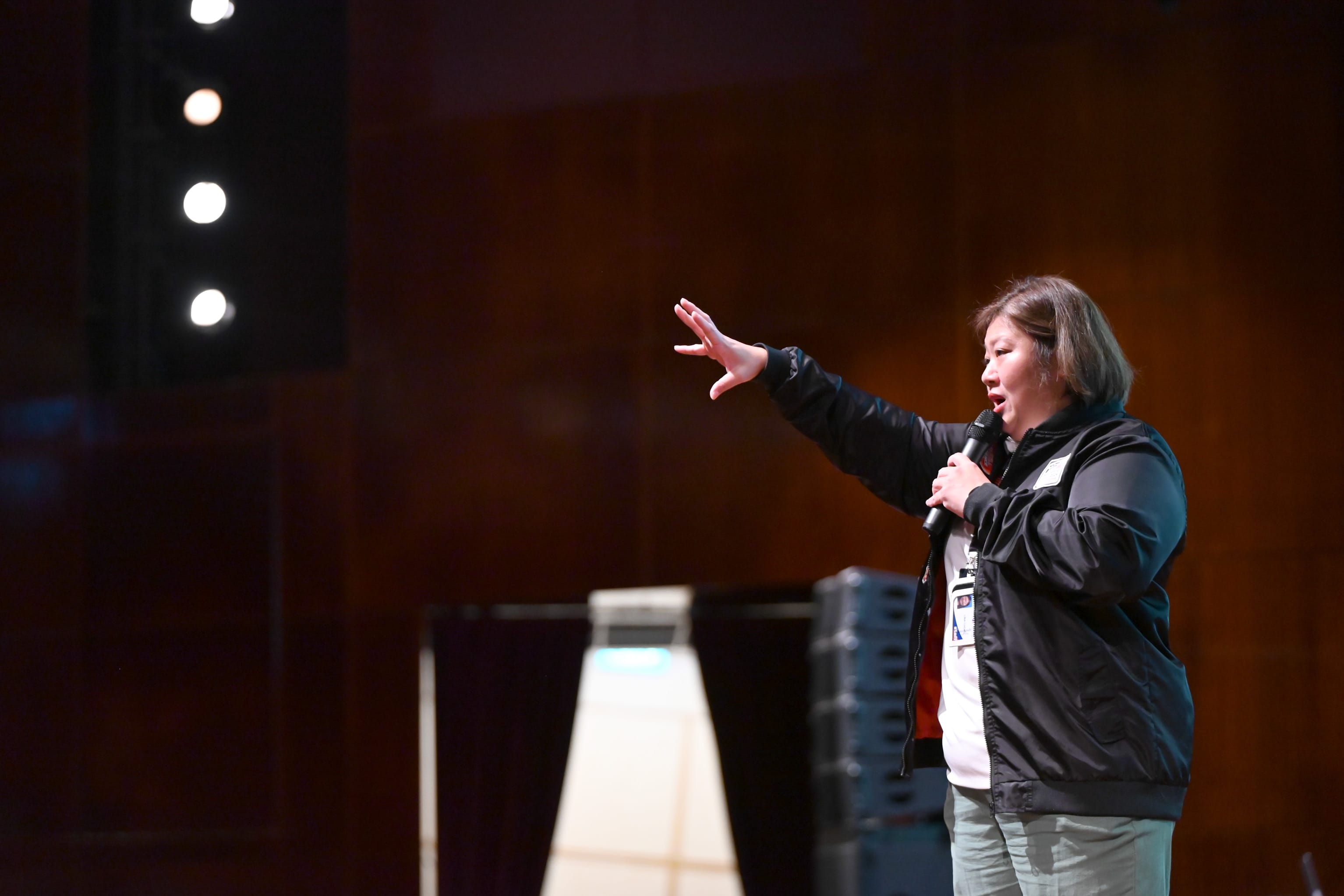 Woman in a black jacket speaks into a microphone on a dimly lit stage, gesturing with one raised hand.