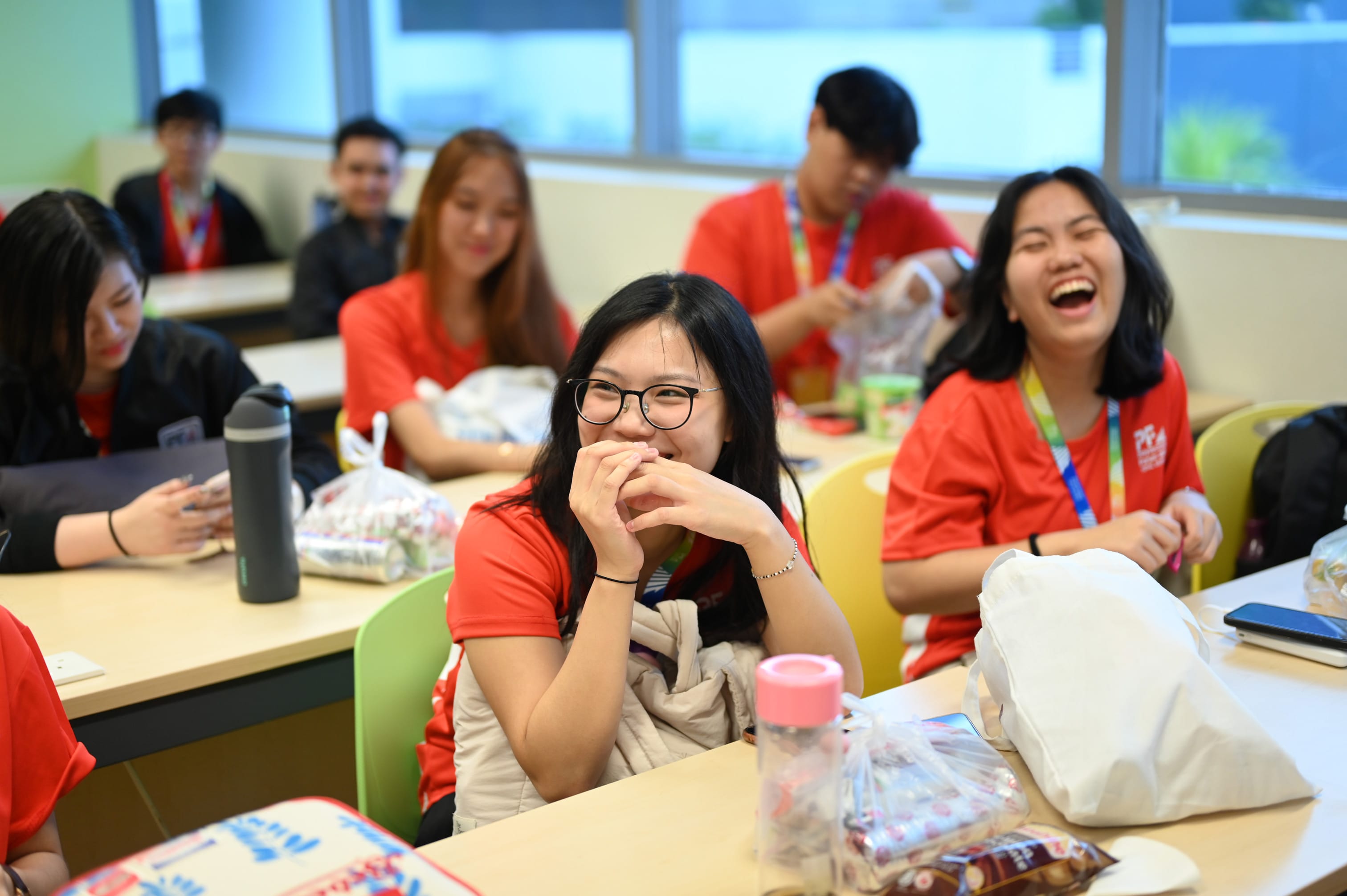 Young people in red shirts sit at classroom desks, smiling and laughing, with snacks and bags on the tables.