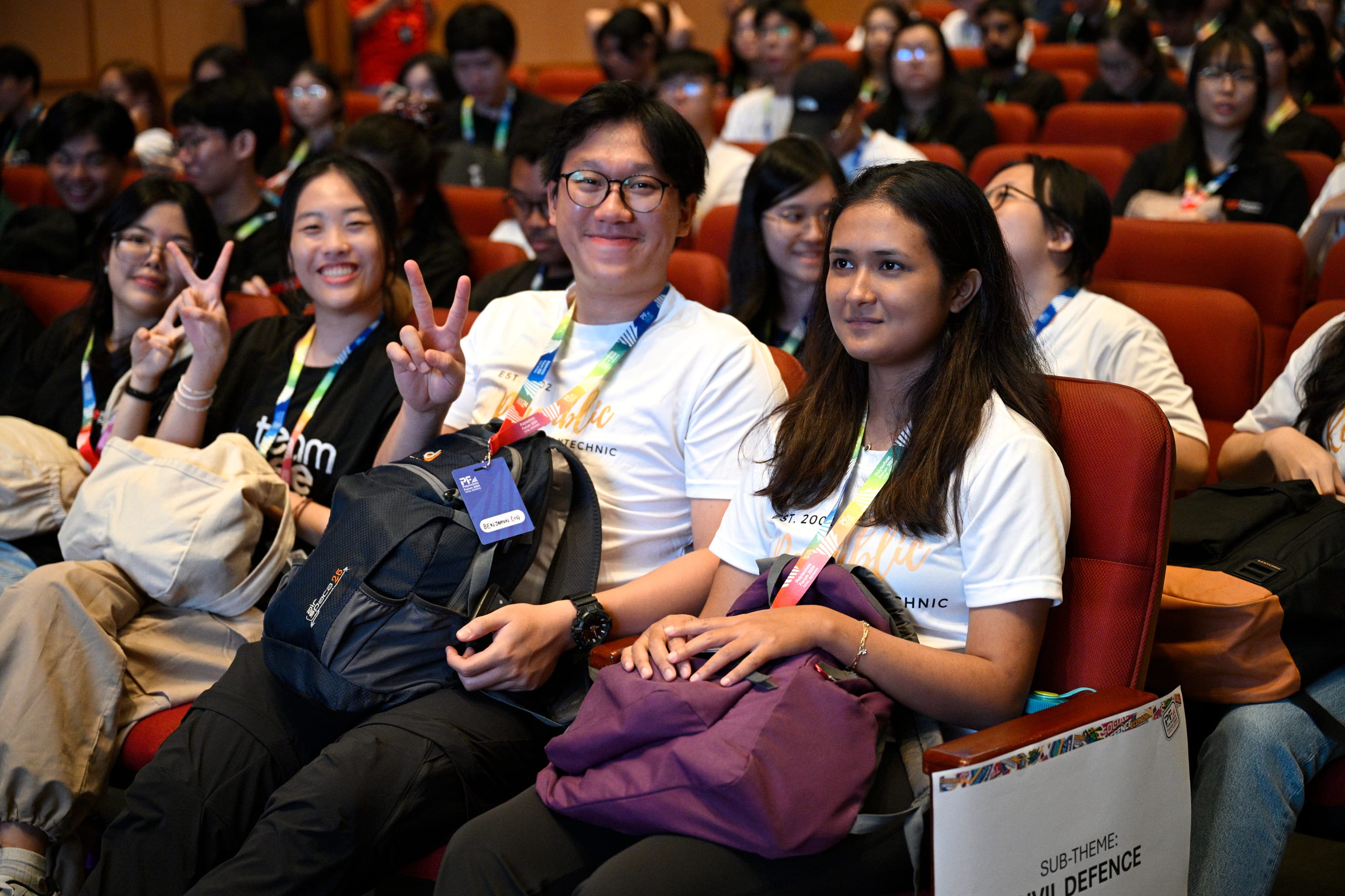 Young people wearing lanyards sit in red auditorium seats, smiling at the camera; two make peace signs.