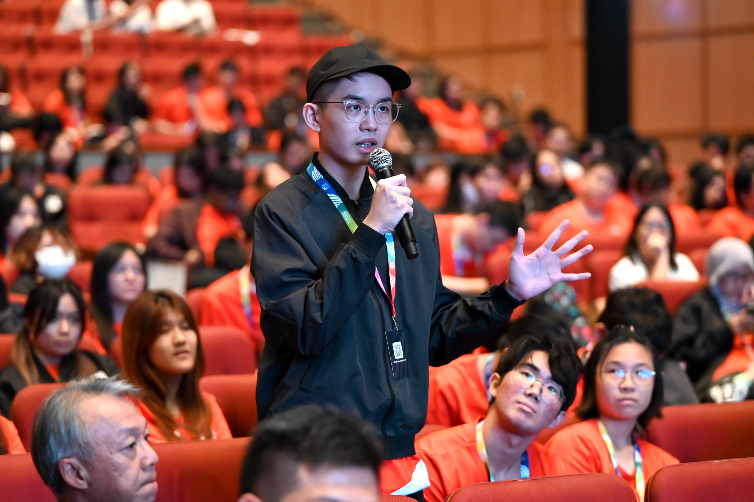 Young man in black cap and jacket speaks into a microphone while gesturing, surrounded by a seated audience in red chairs.