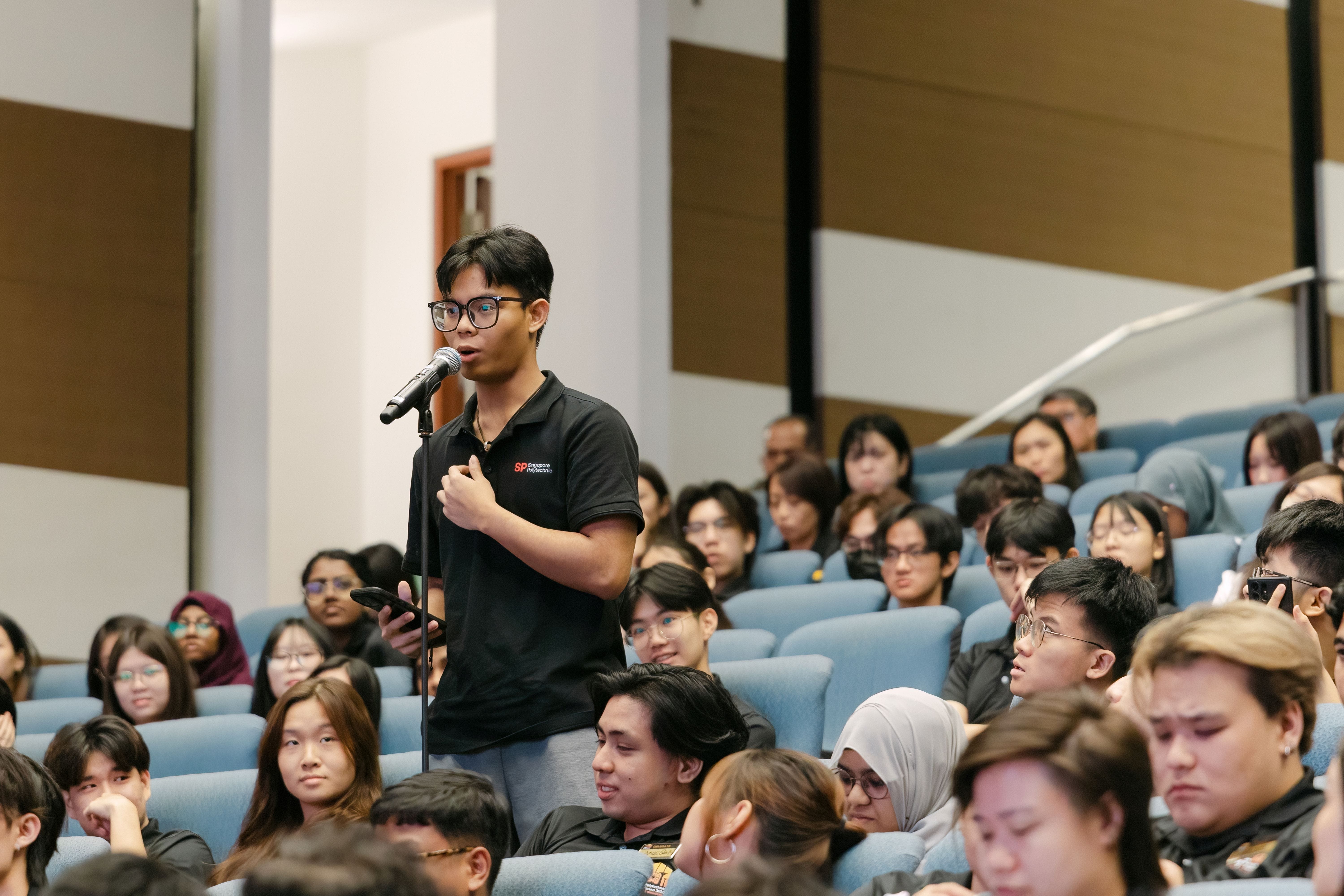 A young man wearing glasses speaks into a microphone while standing among a large seated audience in an auditorium.