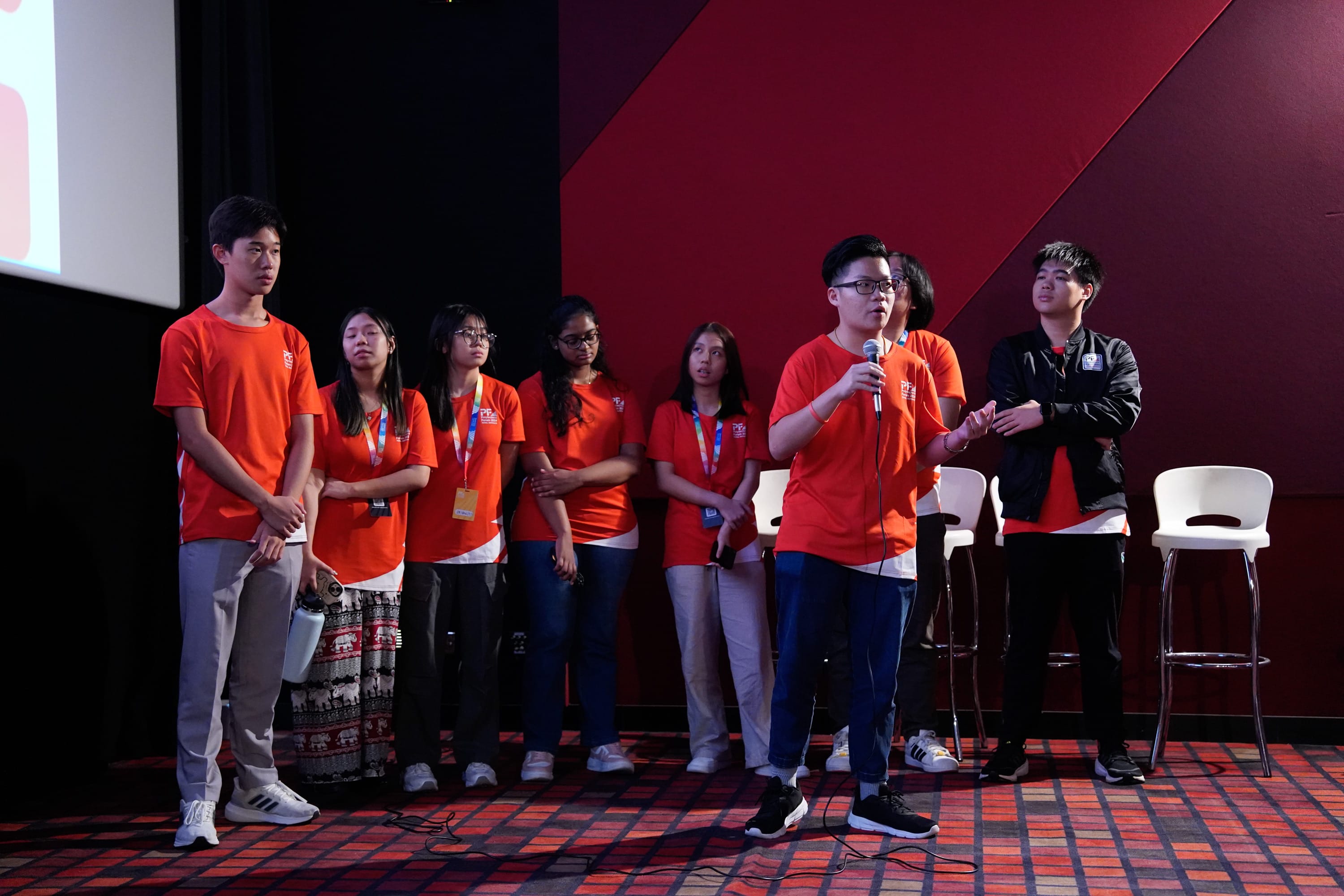 A group of young people in orange shirts stand on a stage while one speaks into a microphone.