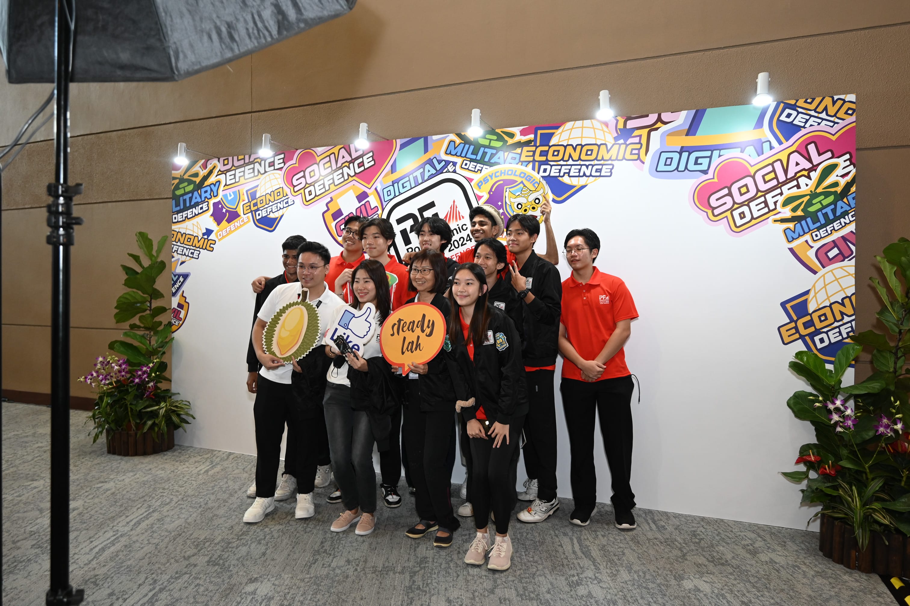 A group of young students posing in front of a colorful defence-themed backdrop, holding fun props including a durian cutout.